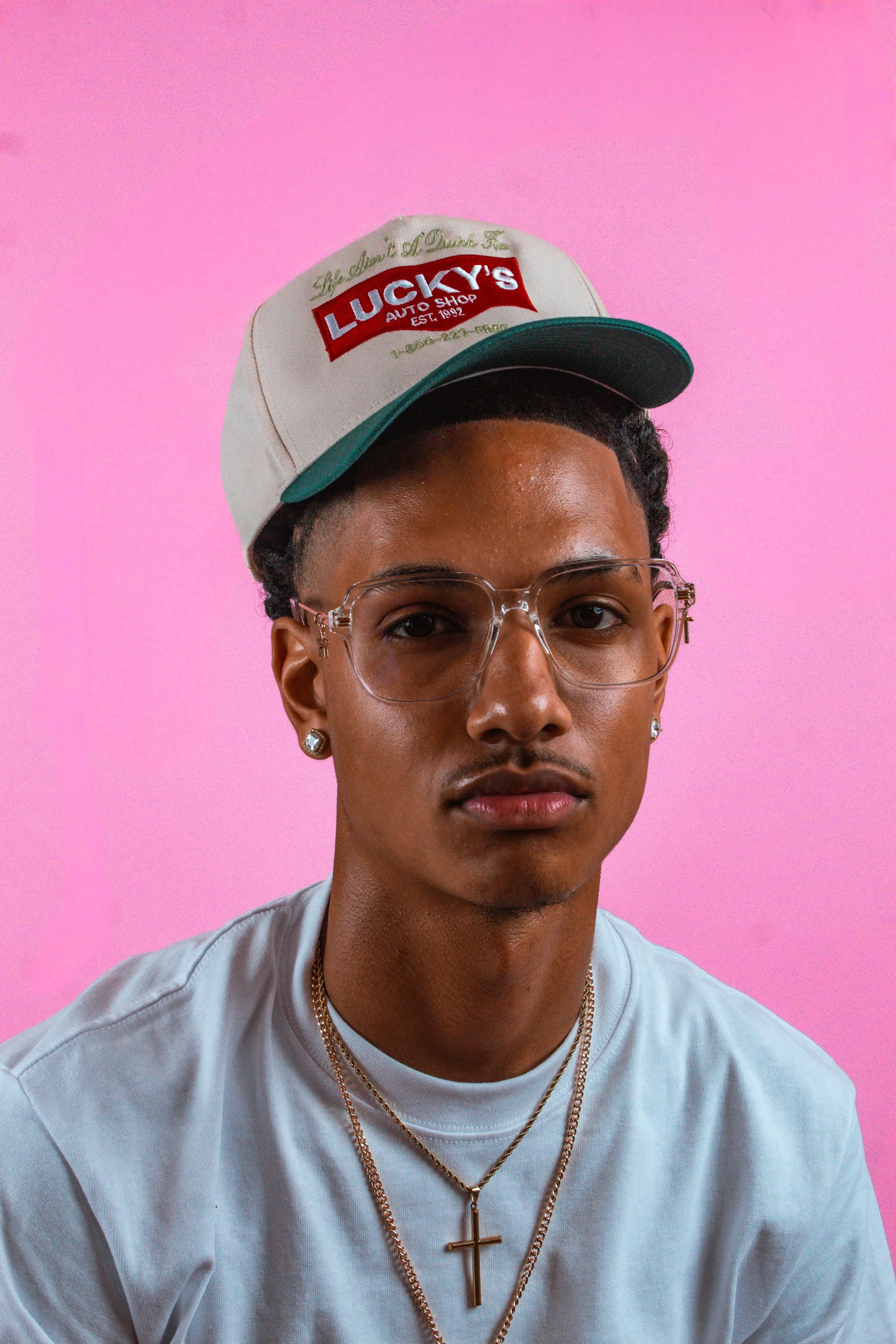 Portrait of a young man wearing glasses, a baseball cap, and jewelry, against a pink background.