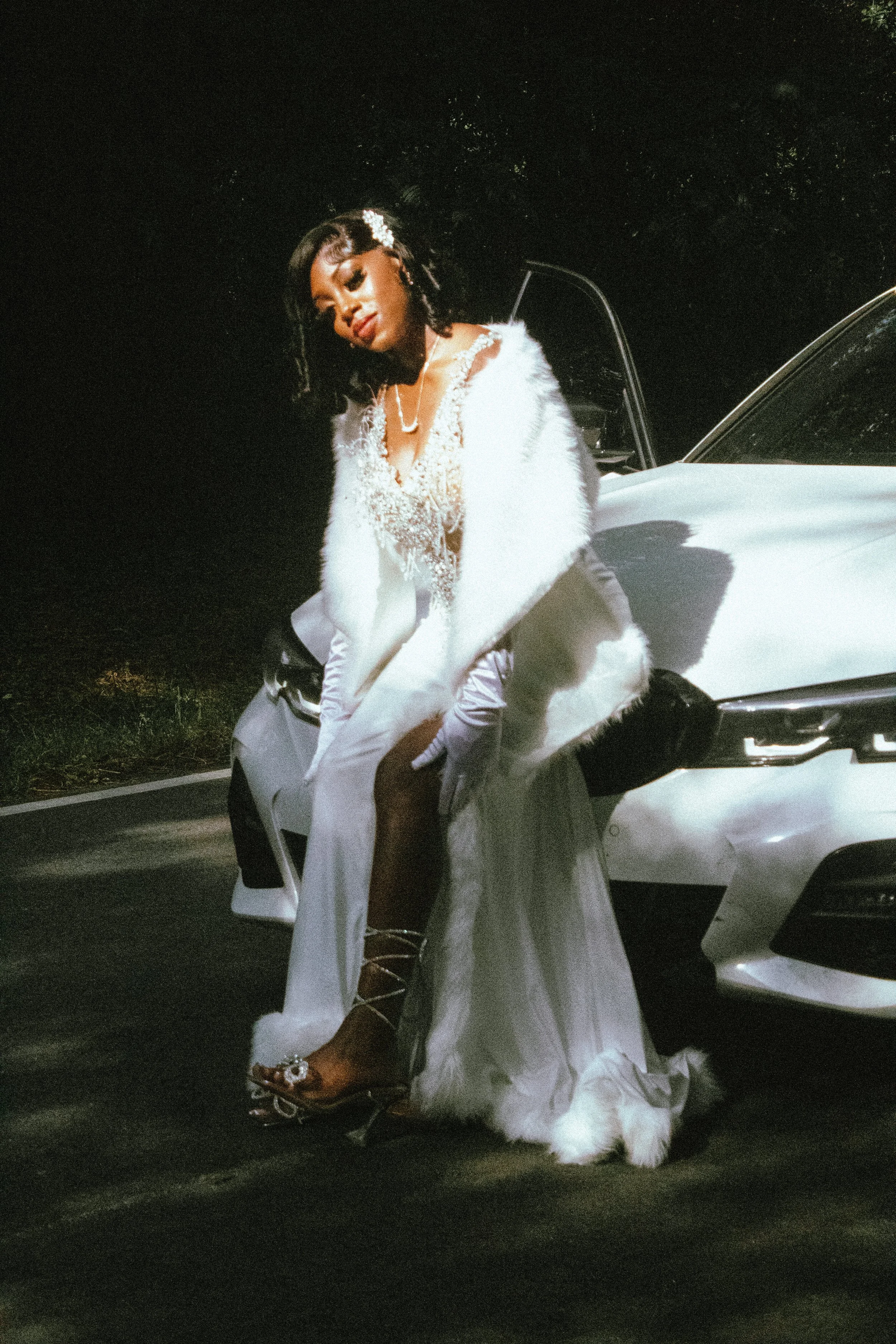 A woman dressed in a white wedding gown and feathered cape, sitting on the front bumper of a white car at night, with a dark background and trees.