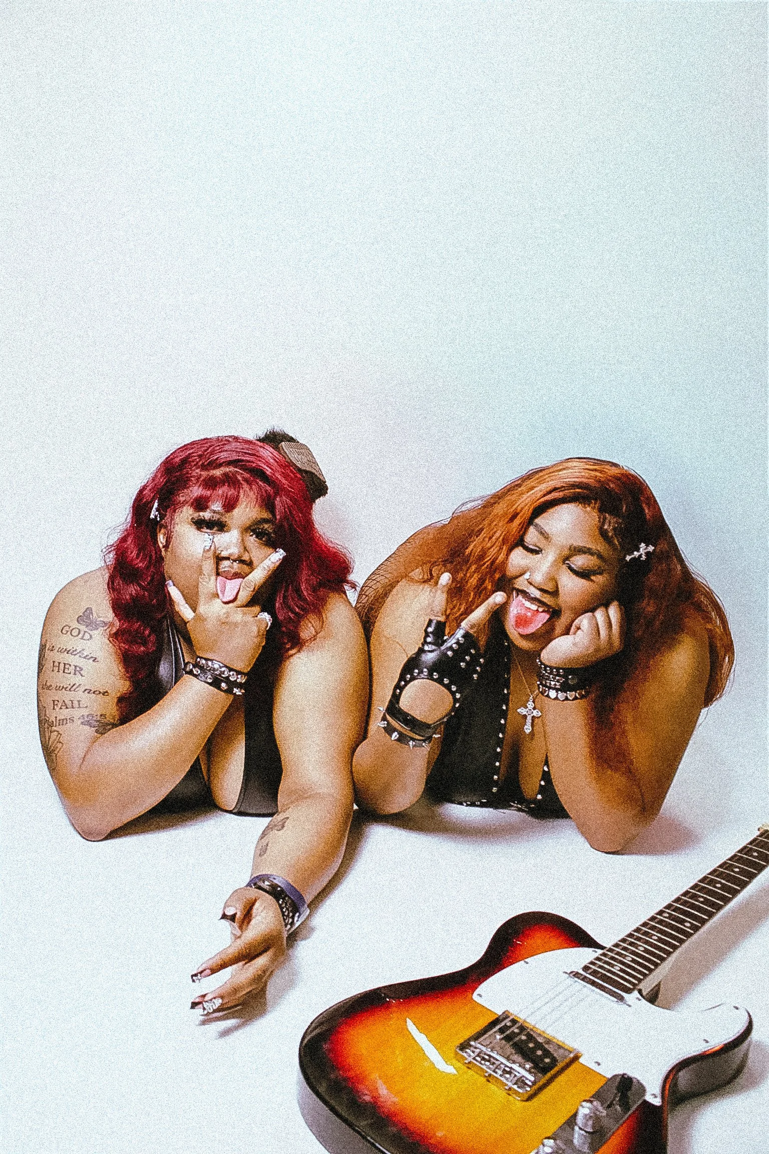 Two women with colorful hair (red and orange), tattoos, and jewelry are lying on a white surface, making peace signs and sticking their tongues out. A sunburst-colored guitar with a black pickguard is lying below them.