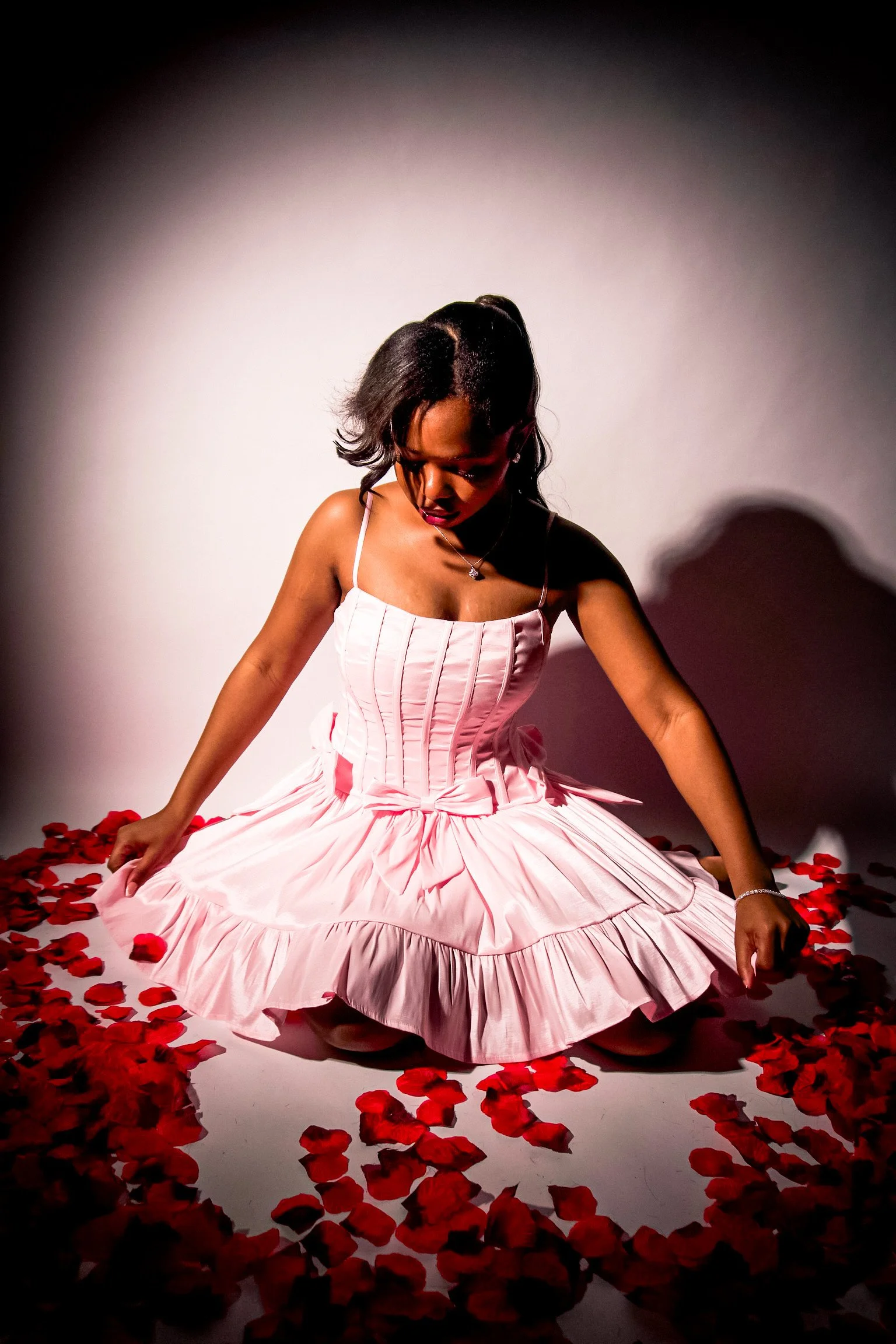 A woman in a pink, pleated ball gown sitting on the floor surrounded by scattered red rose petals.