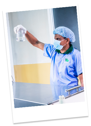 A healthcare worker wearing a blue scrub uniform, face mask, and hairnet holding a glass of clear liquid in a hospital or medical facility.