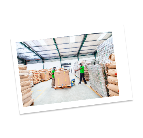 Two workers in green shirts and masks in a warehouse, moving pallets of large paper bags.