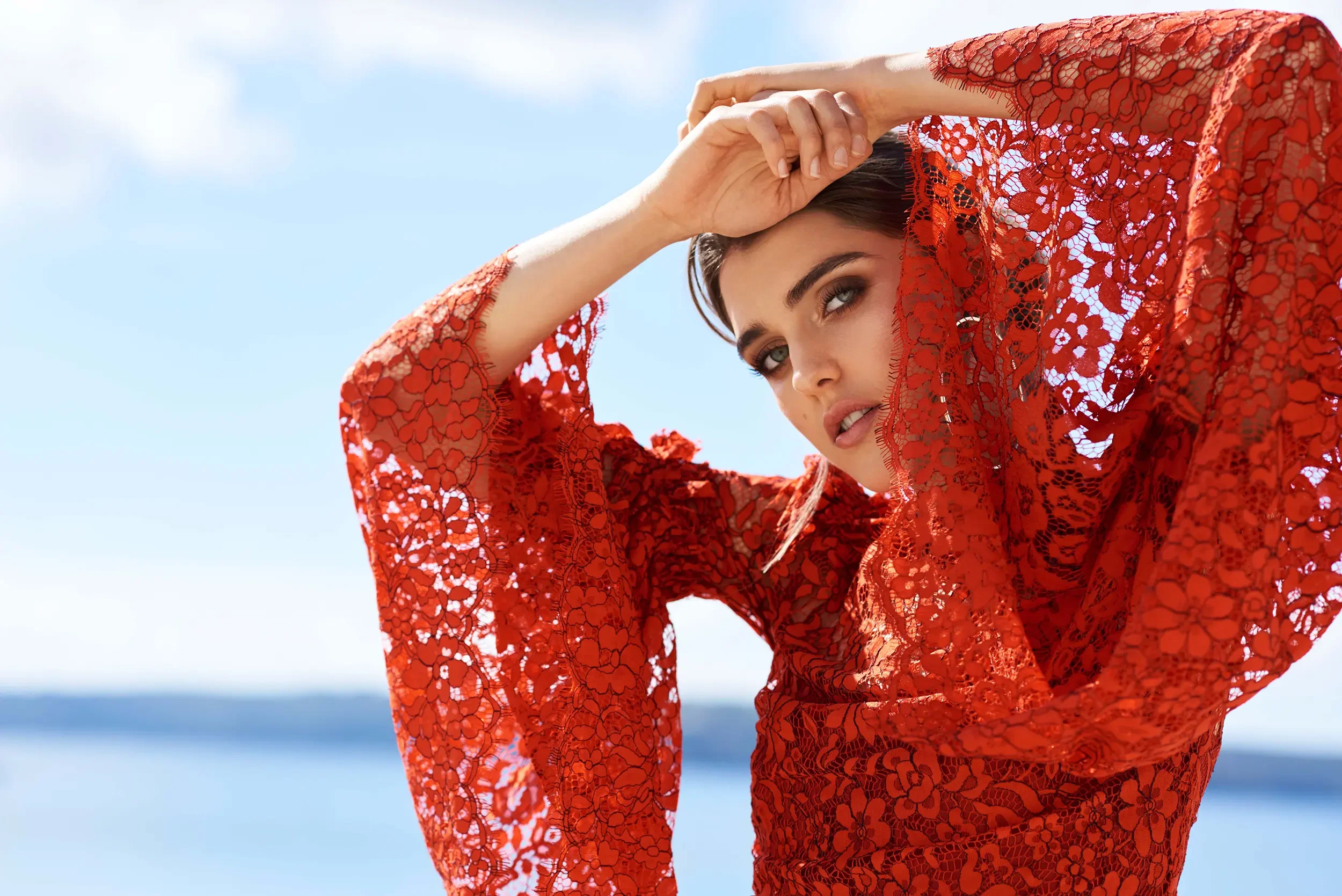 Woman in red lace dress looking at the camera with sky and ocean in the background.