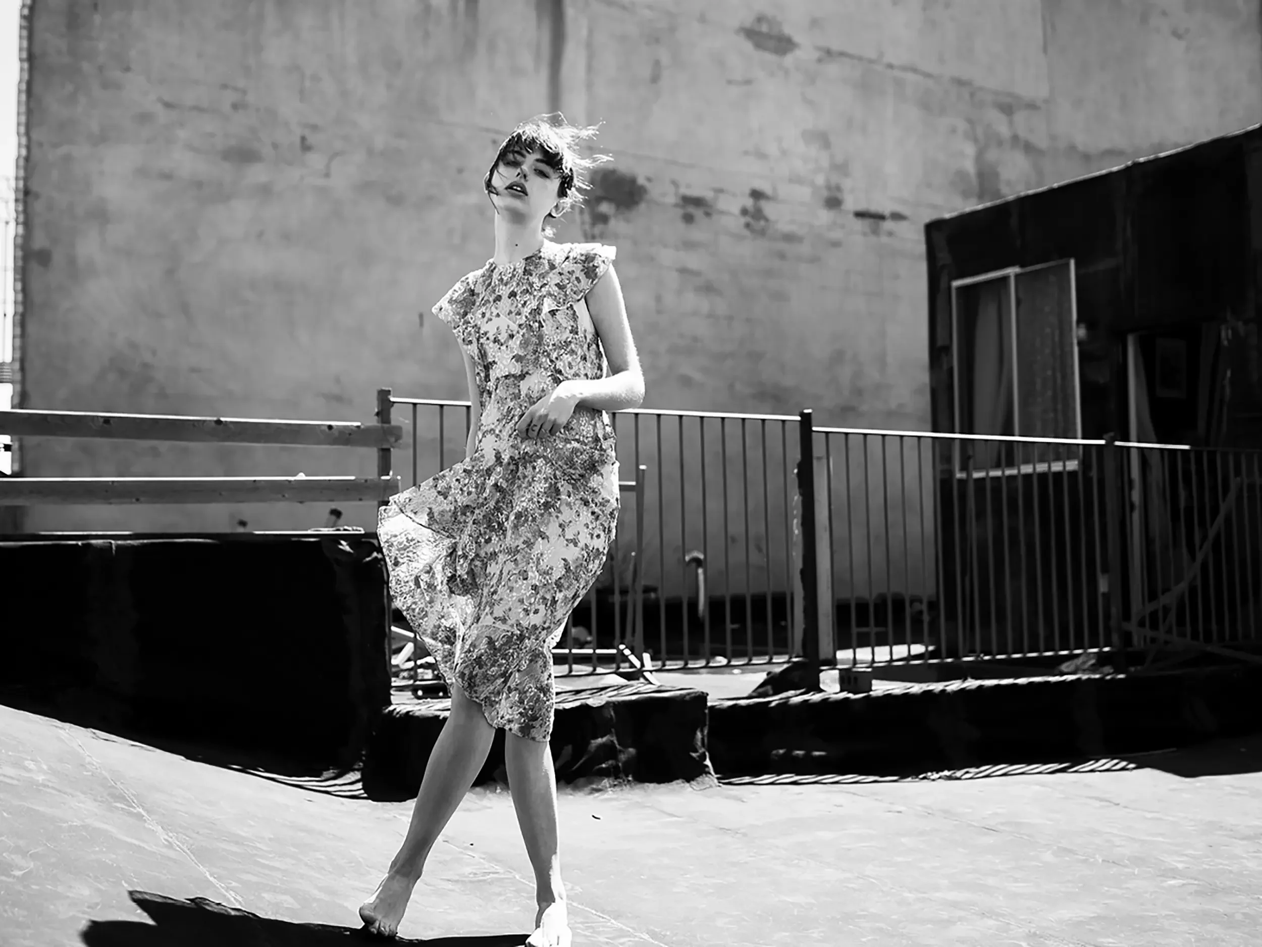 A woman in a floral dress dancing on an outdoor stage with a concrete wall and metal railings in the background, captured in black and white.