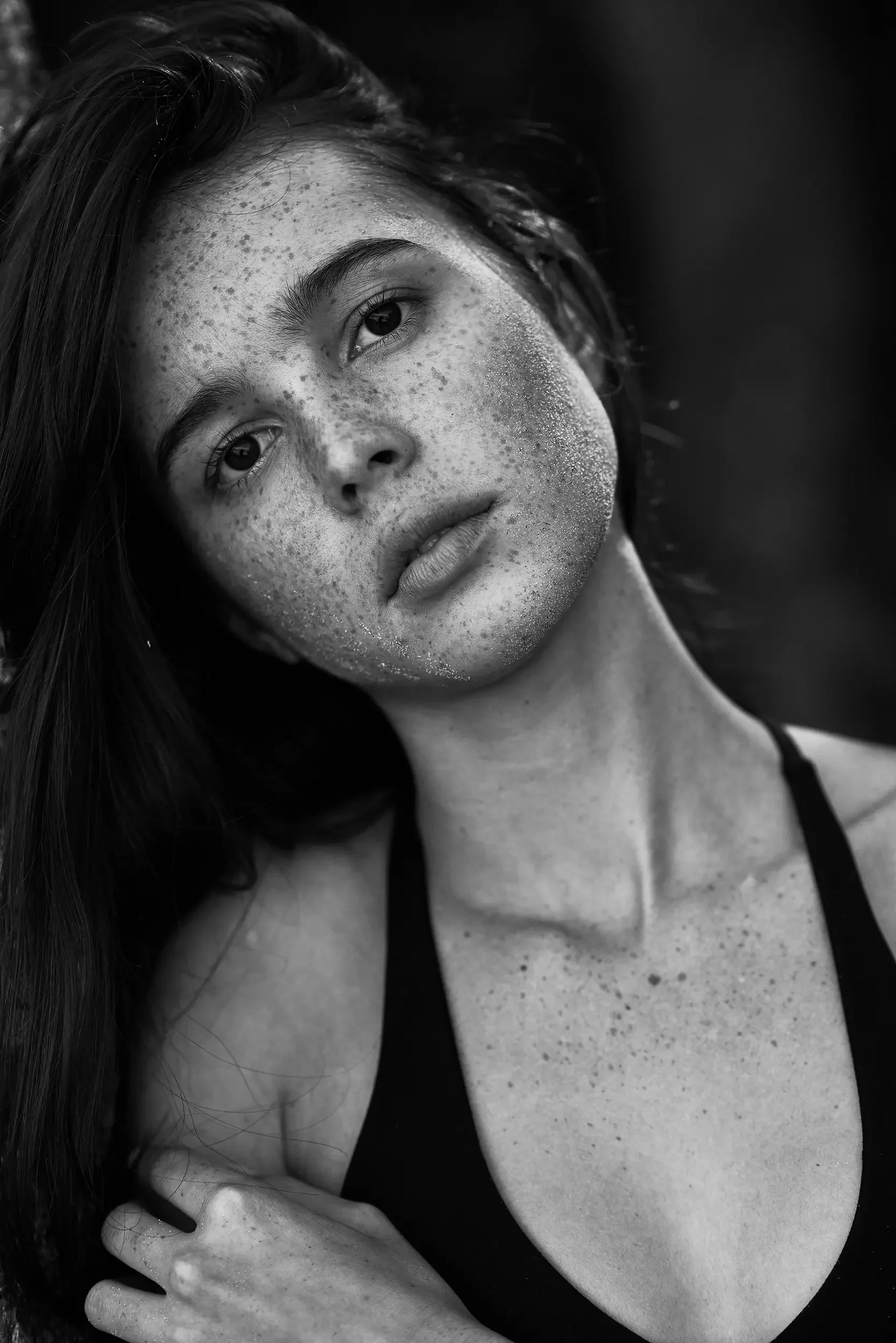 A close-up black-and-white photograph of a woman with wet hair and freckles, gazing softly at the camera with her hand resting on her shoulder, wearing a black sleeveless top.