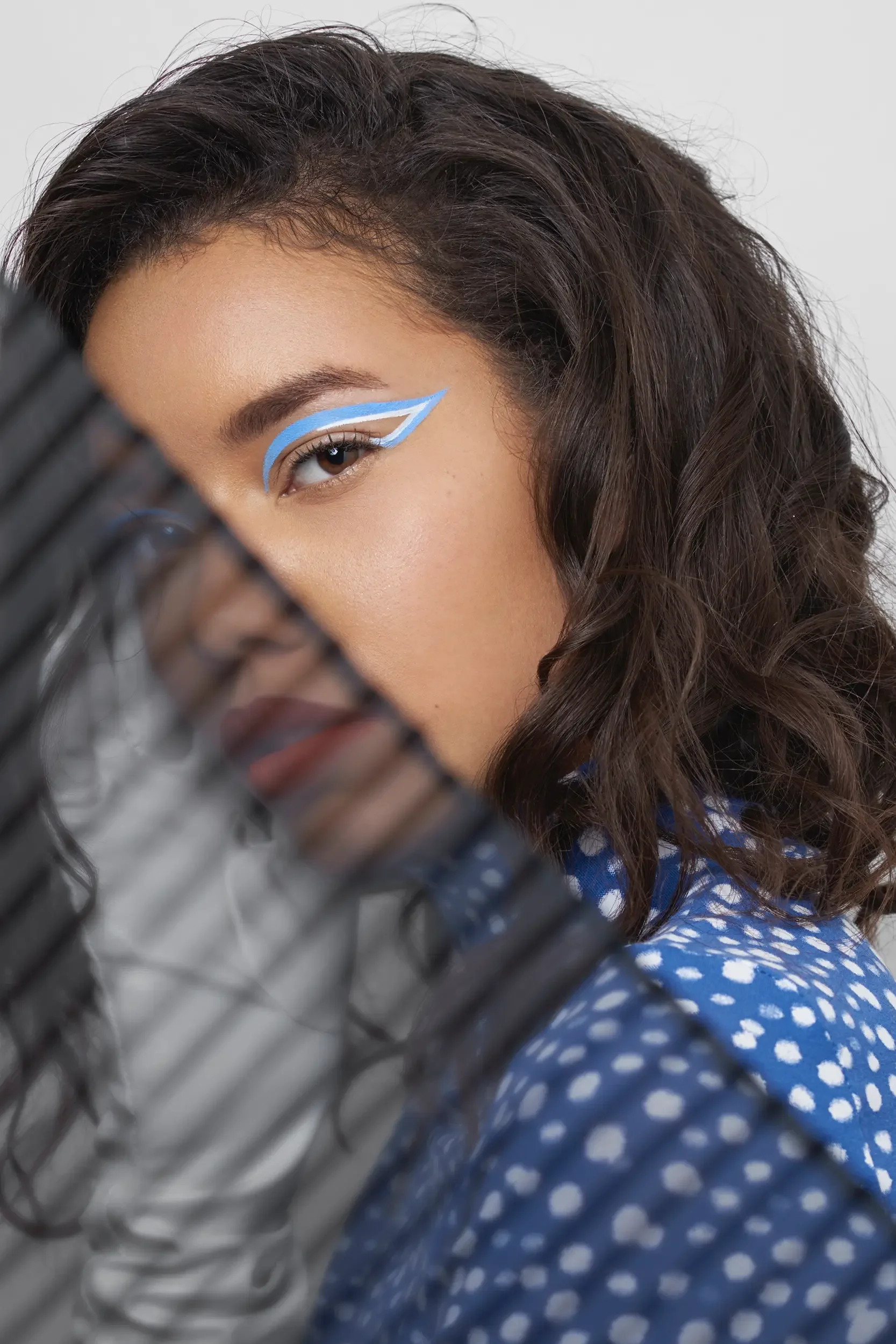 Close-up of a woman's face with curly brown hair and blue eye makeup. She is partially obscured by a black grill or grate.