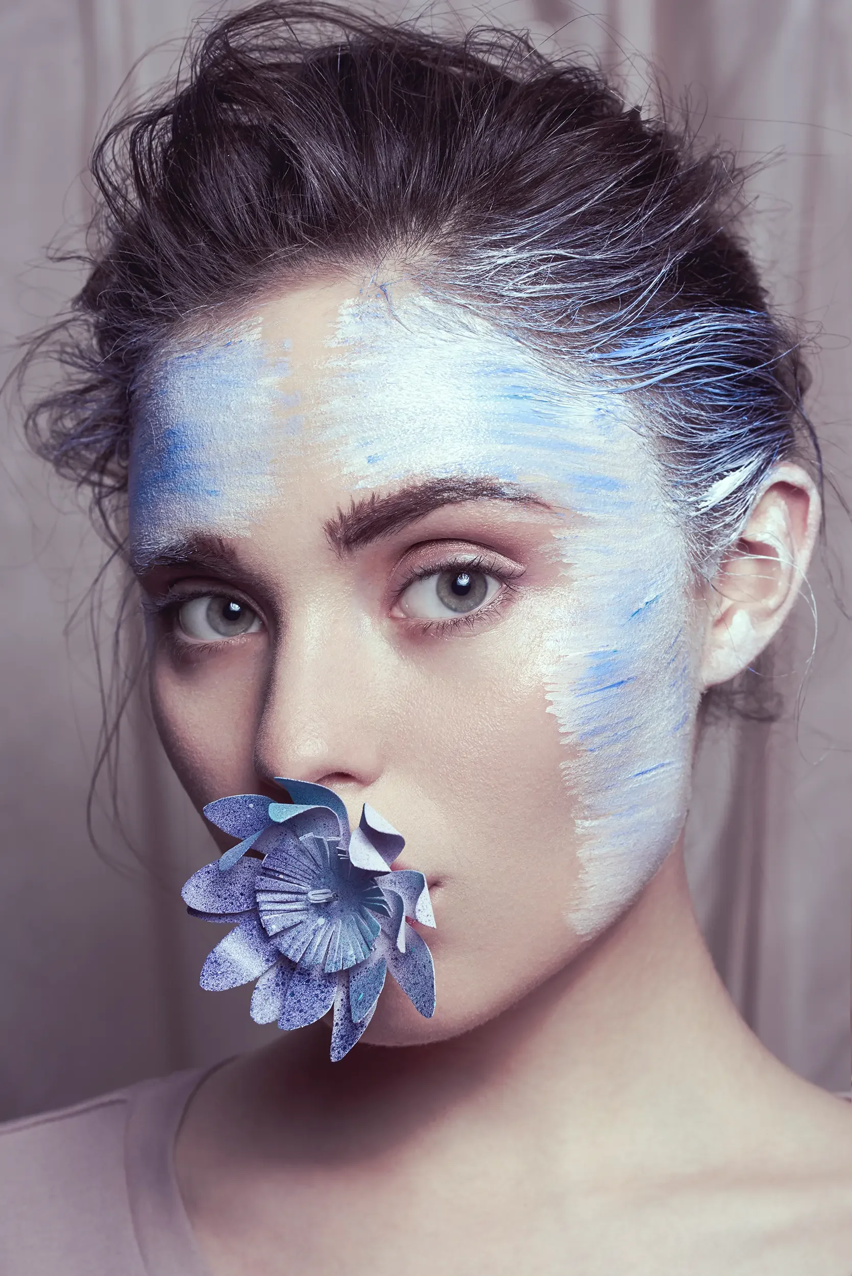 A woman with gray eyes and dark hair, with her face painted in shades of white and blue, holding purple flowers near her face.