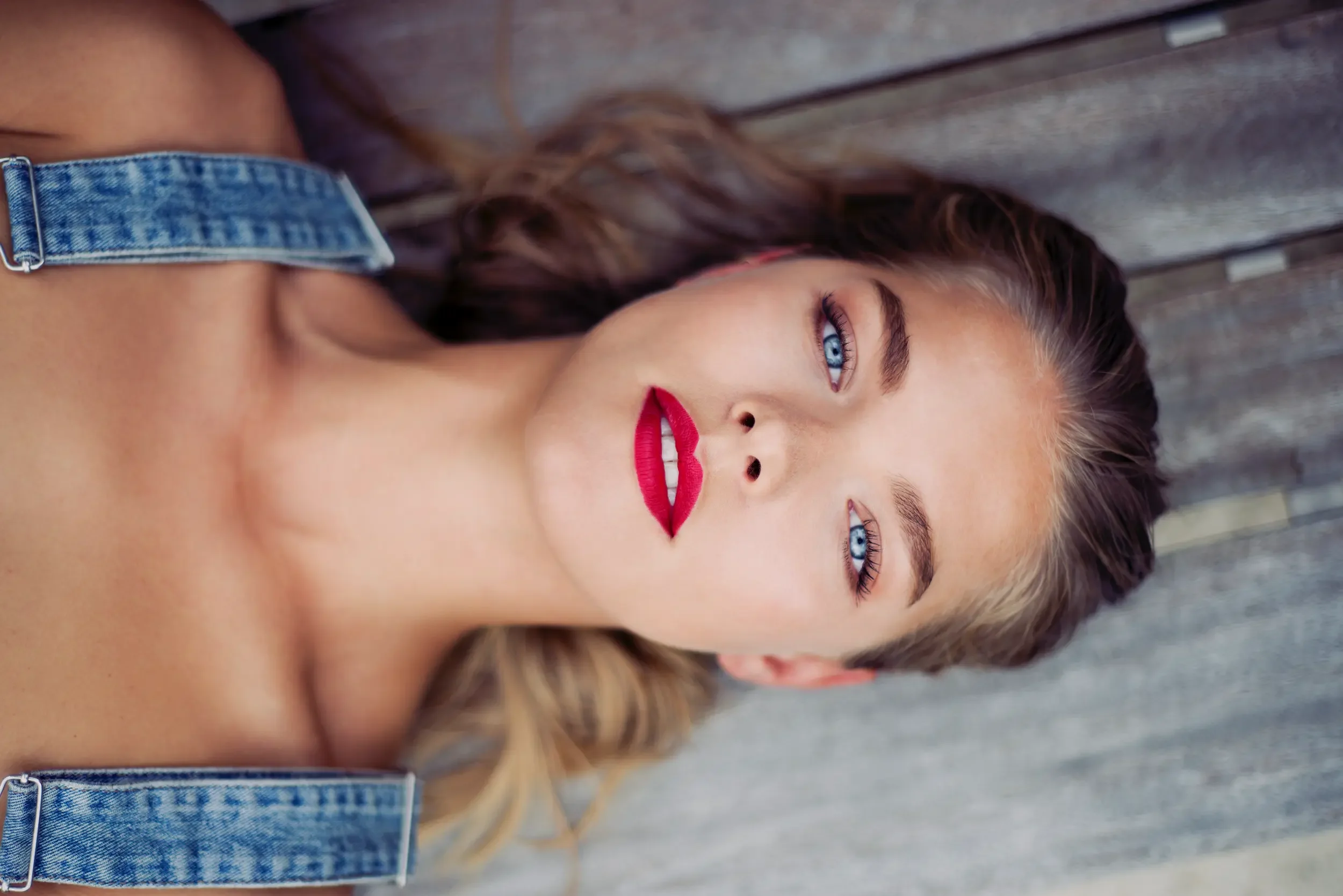 A woman with blue eyes, red lipstick, and wavy brown hair laying on a wooden surface, wearing a denim strap top.