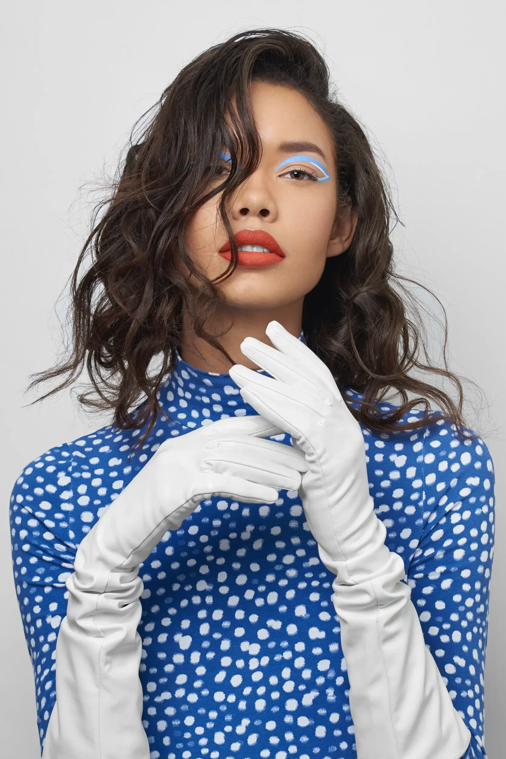 A woman with wavy brown hair, wearing a blue polka-dot dress and white gloves, touching her face with her left hand while her right is near her neck, showcasing bold makeup and expressive pose.