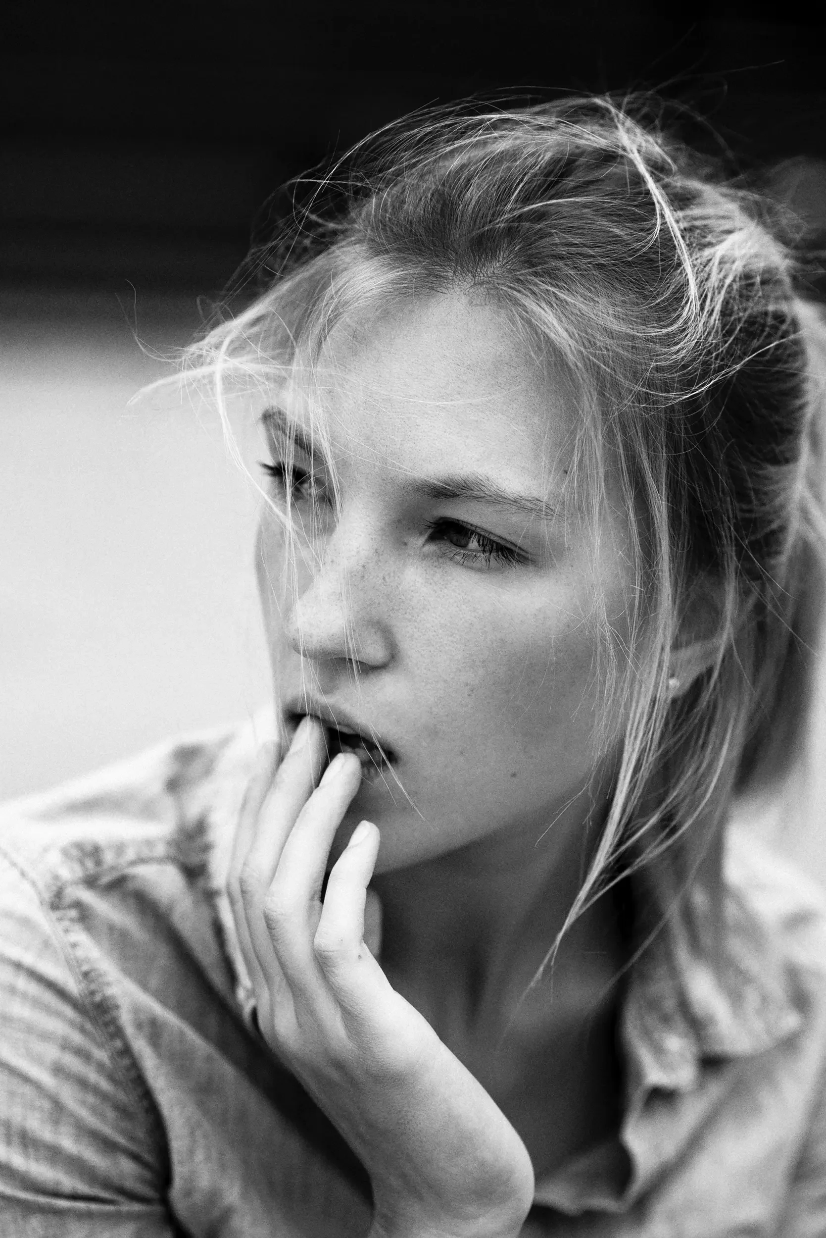 Black and white close-up of a young woman's face with her hand gently touching her chin, visible freckles, light-colored hair, and a contemplative expression.