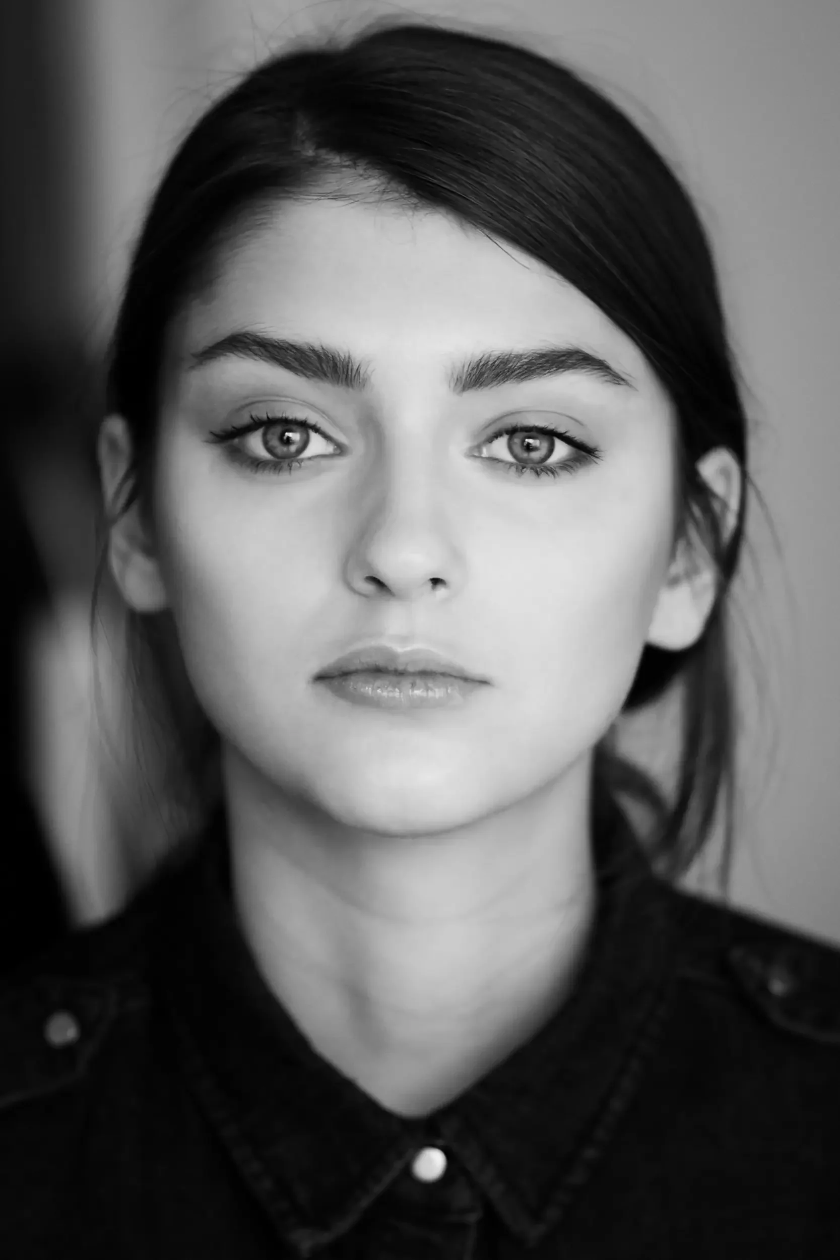 Black and white close-up portrait of a young woman with long dark hair, piercing eyes, and wearing a dark jacket with buttons.