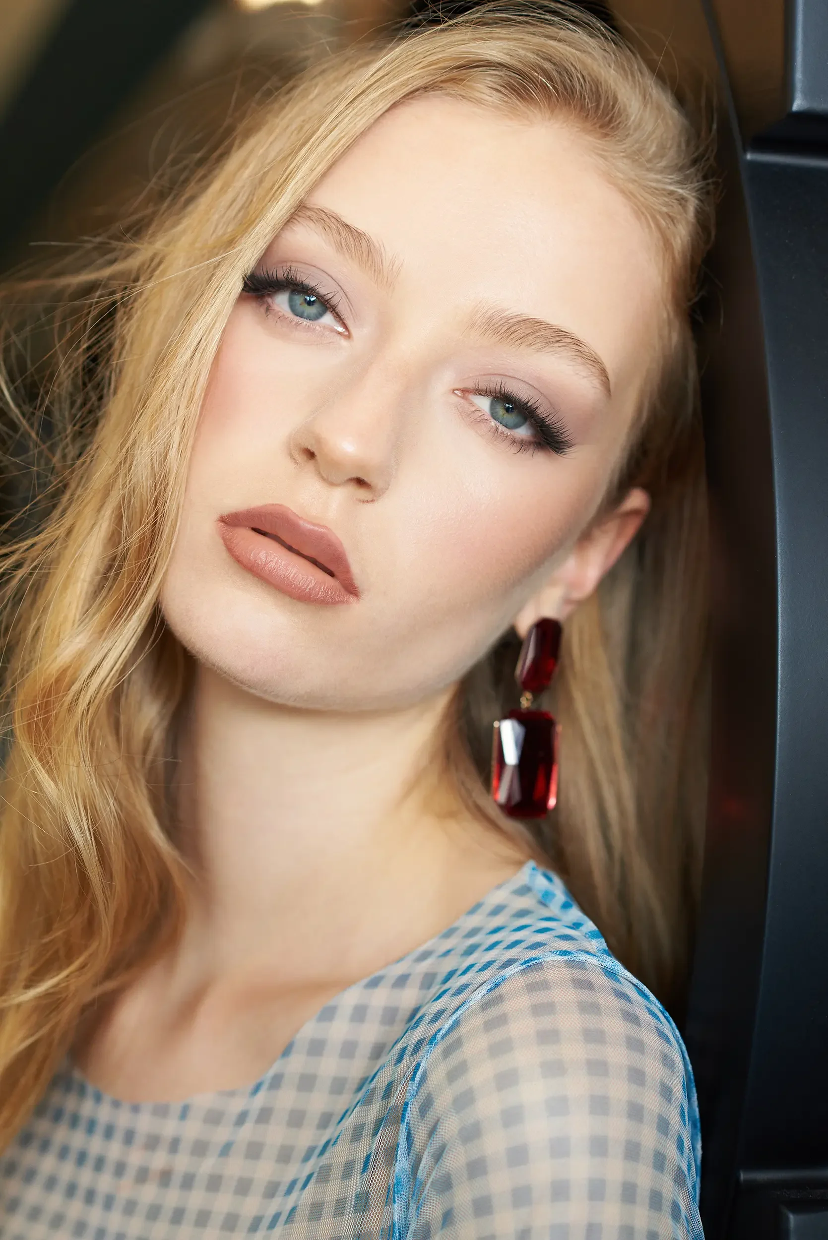 Close-up of a young woman with blonde hair and blue eyes, wearing makeup and red earrings, looking at the camera.