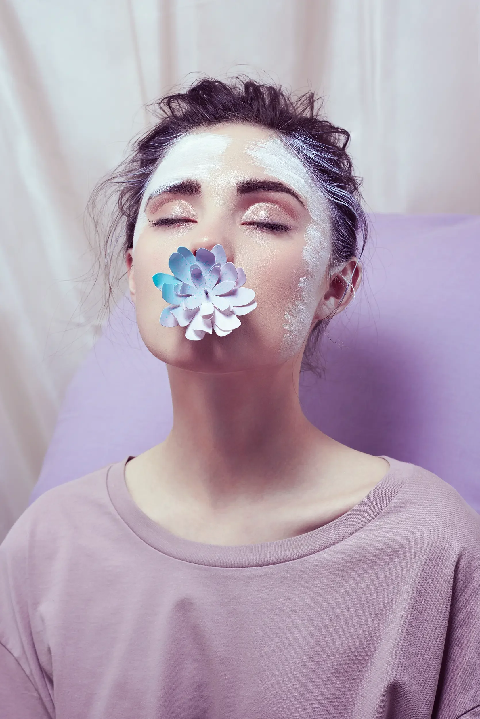 A woman with dark hair and closed eyes receiving facial treatment, with a white facial mask and a blue and white flower on her nose, wearing a pink shirt, against a lavender background.