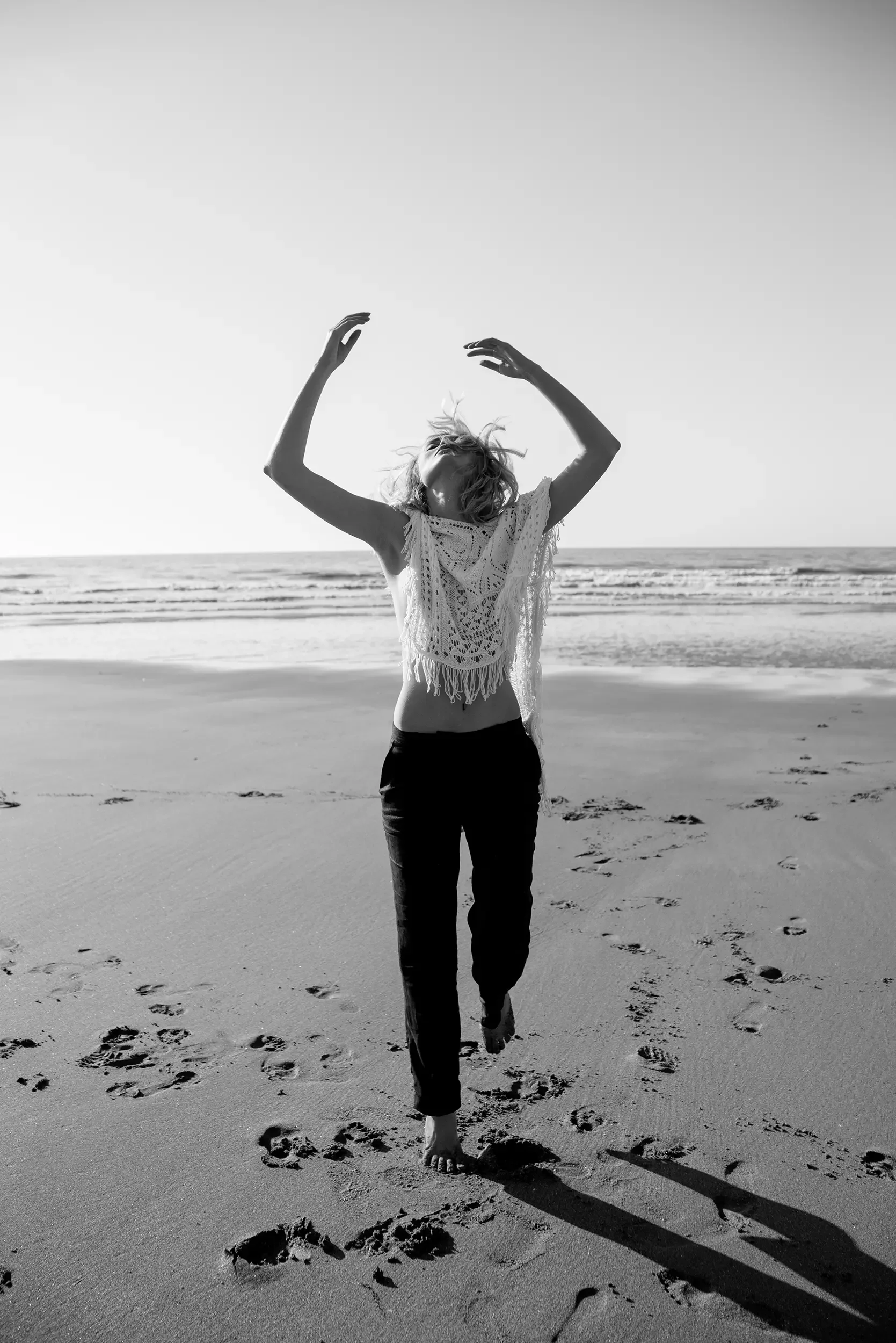 A woman with curly hair walking barefoot on a sandy beach during sunset, with the ocean in the background, wearing a lace top. The photo is black and white.
