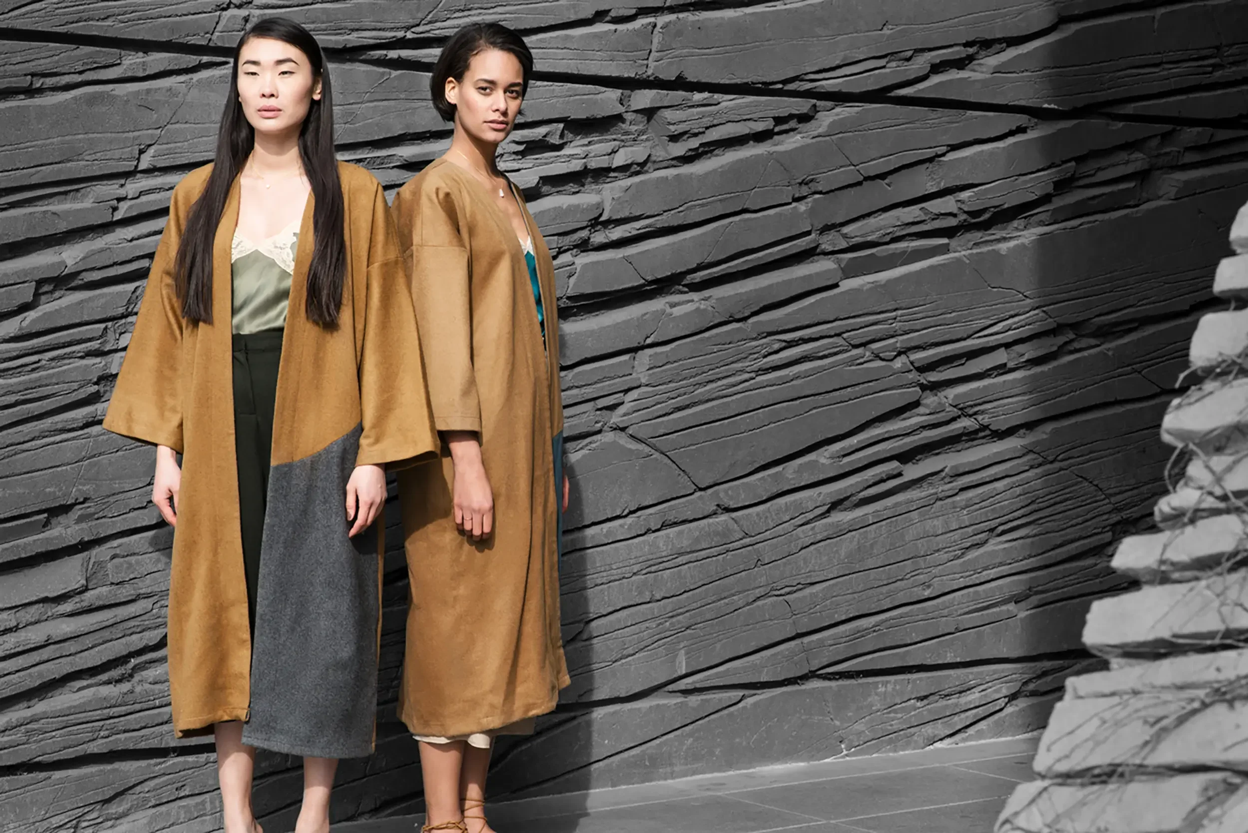 Two women standing side by side in front of a dark stone wall, wearing long brown coats and stylish dresses.