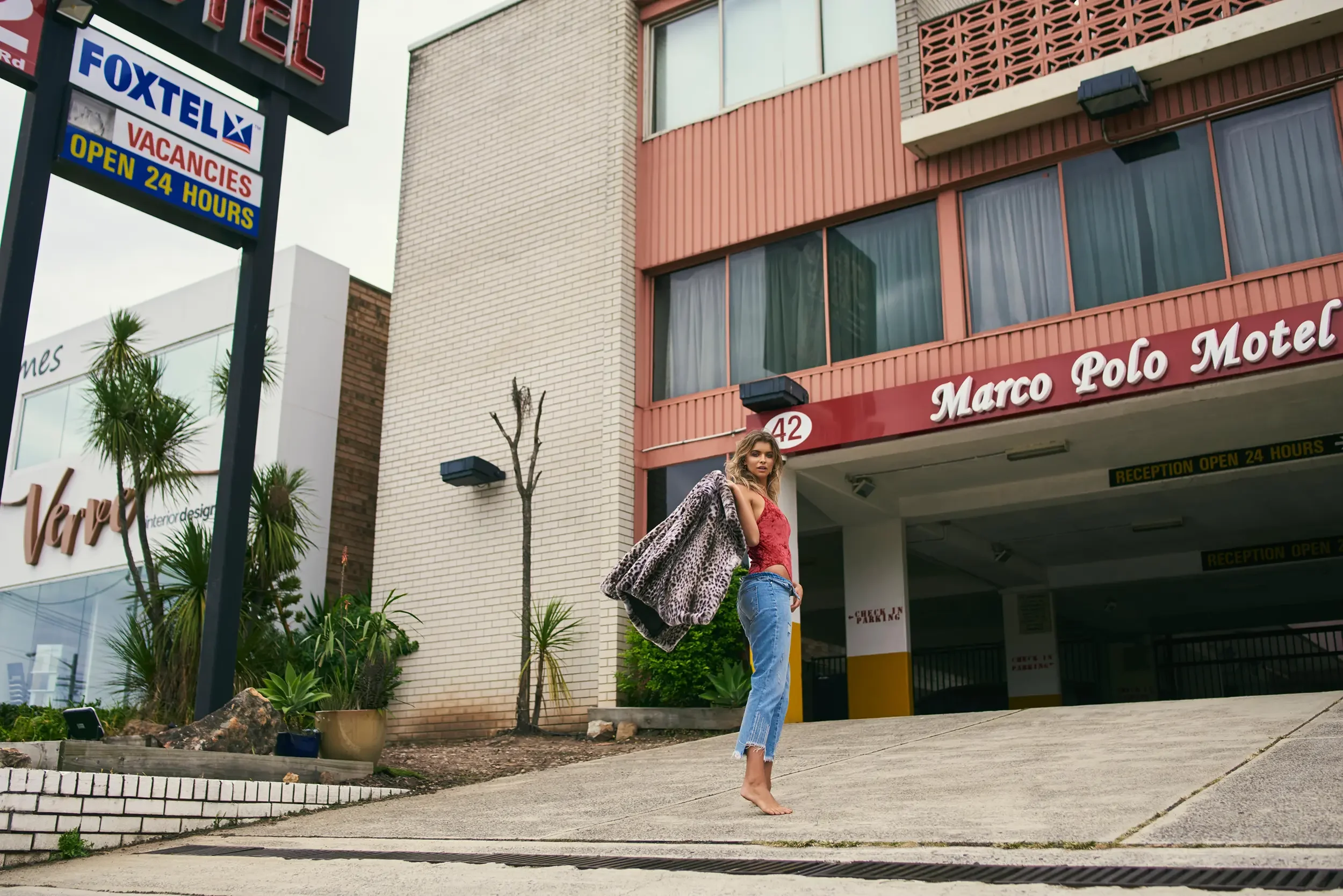 A young woman in a red top, blue jeans, and a leopard print coat stands without shoes in front of a motel building, with signs indicating vacancies and 24-hour reception. There are potted plants and trees around her, and the building has pinkish-brow