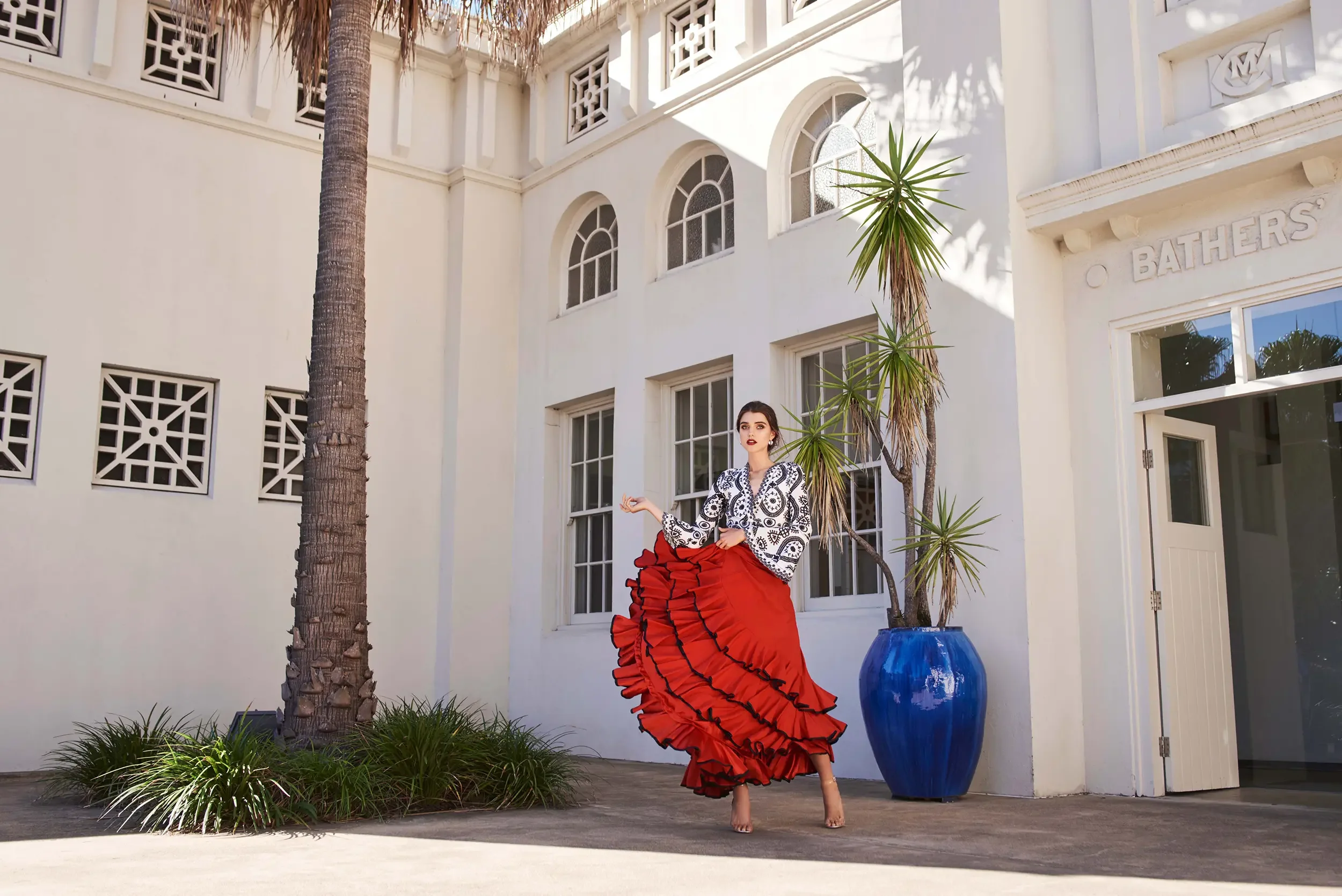 A woman in a black and white patterned blouse and a long red ruffled skirt standing outdoors in front of a white building with large windows, palm trees, and a blue pot.