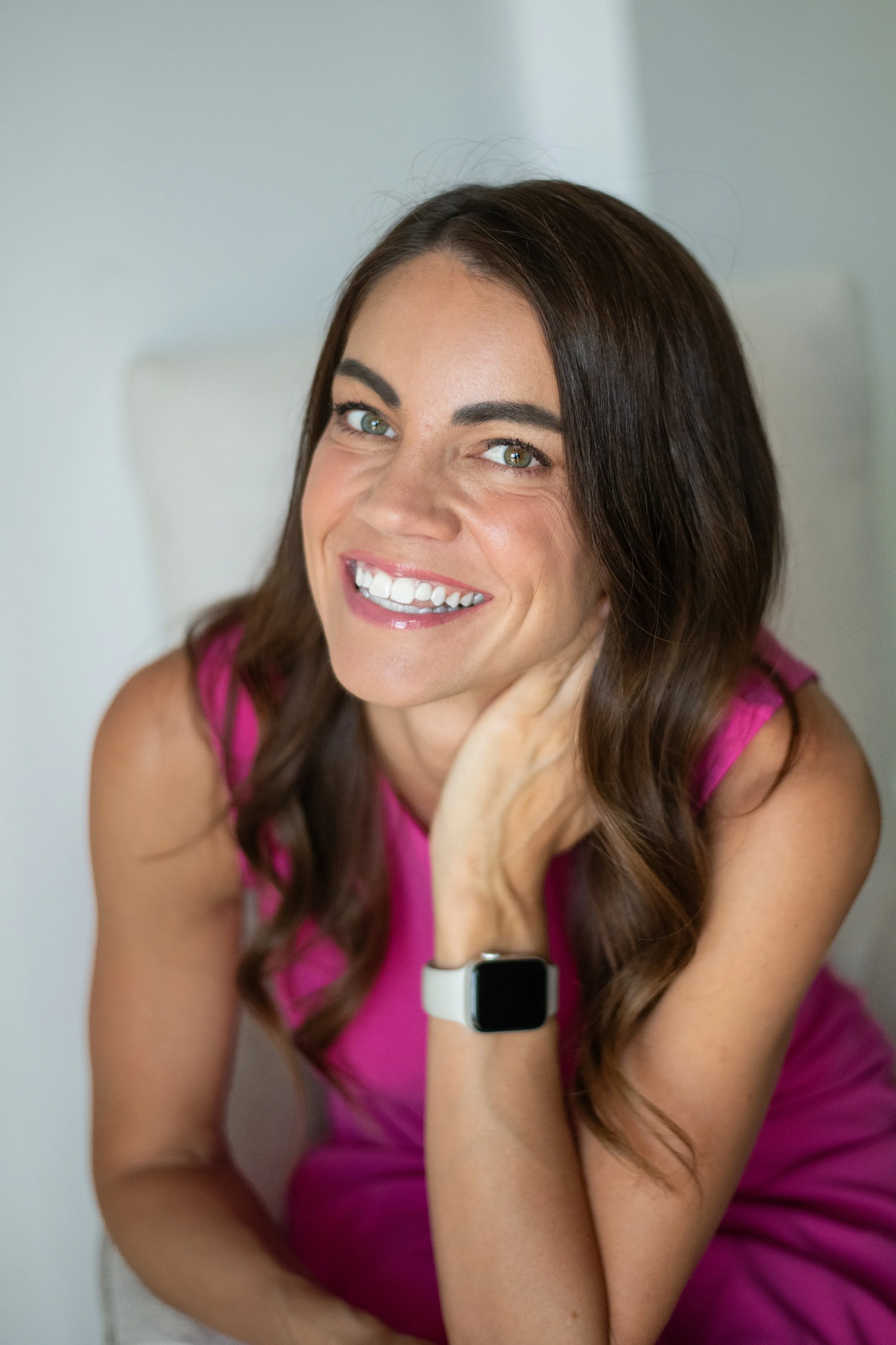 A woman with long brown hair, wearing a pink sleeveless top, smiling and looking at the camera. She is resting her chin on her hand, with a smartwatch on her wrist. The background is plain and light-colored.