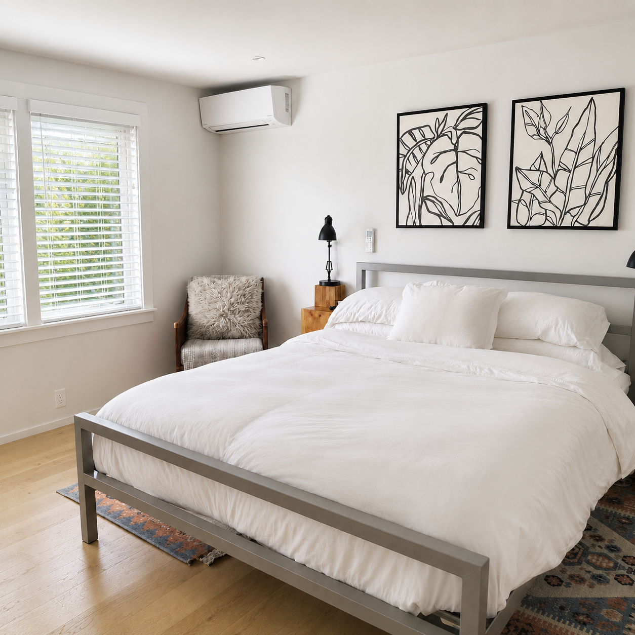 Modern bedroom with white bedding, black and white framed art, wooden side tables, black reading lamp, window with white blinds, and an air conditioning unit.