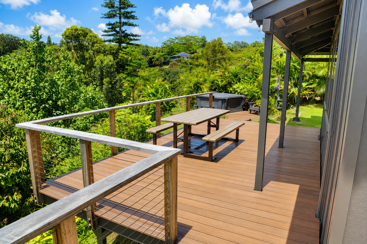 A spacious wooden deck attached to a house, surrounded by lush green trees and plants, with a picnic table and benches, a hot tub, and outdoor lighting on a sunny day.