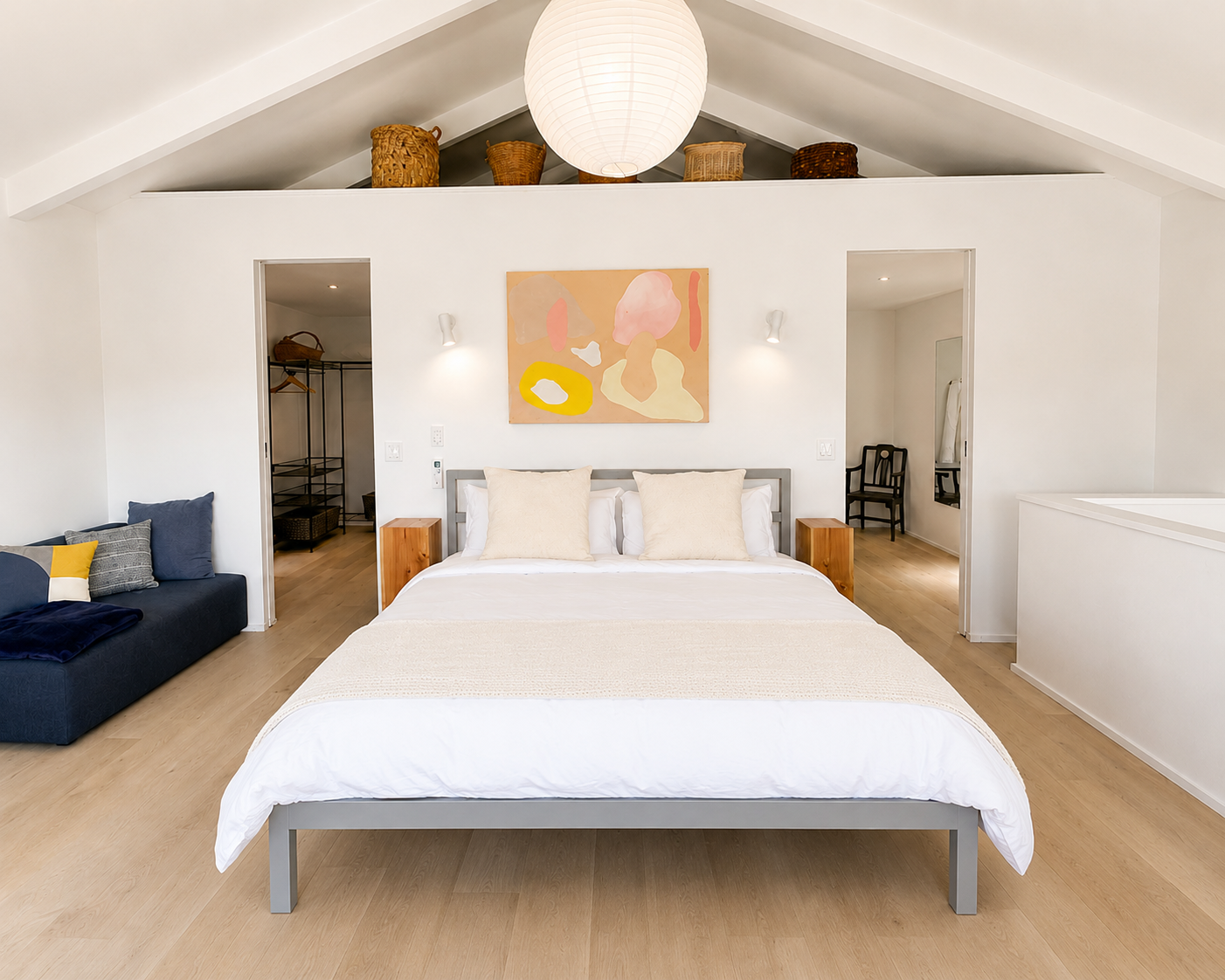 A modern bedroom with a large bed, two wooden nightstands, and abstract art hanging above the headboard. There is a black sofa with cushions to the left and a walk-in closet in the background. A paper lantern hangs from the ceiling, and the room has 