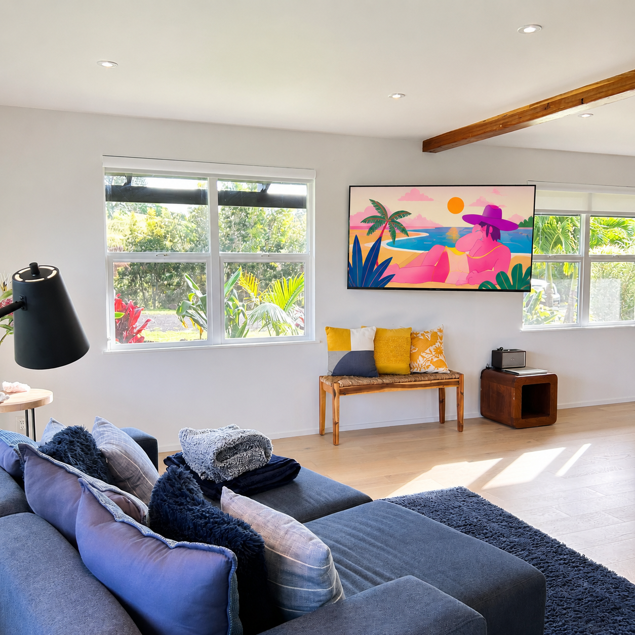 Living room with a gray sectional sofa, blue pillows, a black lamp, a wooden bench with yellow and white cushions, a wall-mounted colorful beach scene artwork, large windows with lush green foliage outside, and a wooden side table with a small speake