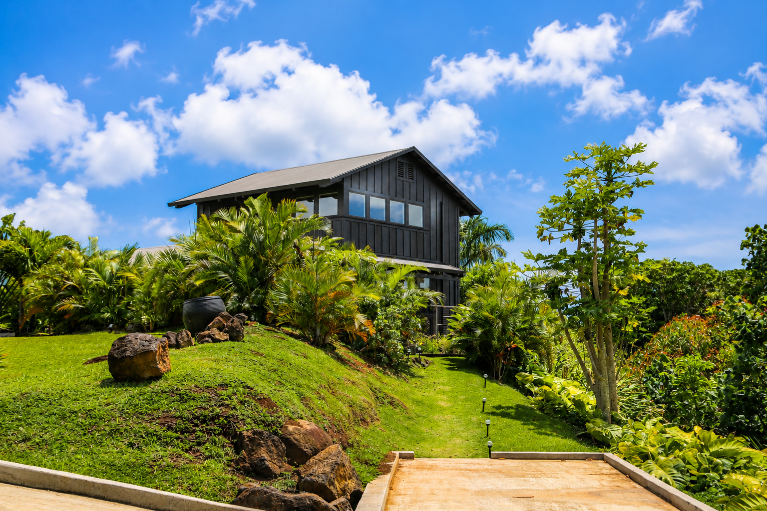 A modern black house on a lush green hillside with tropical plants and a bright blue sky with white clouds.