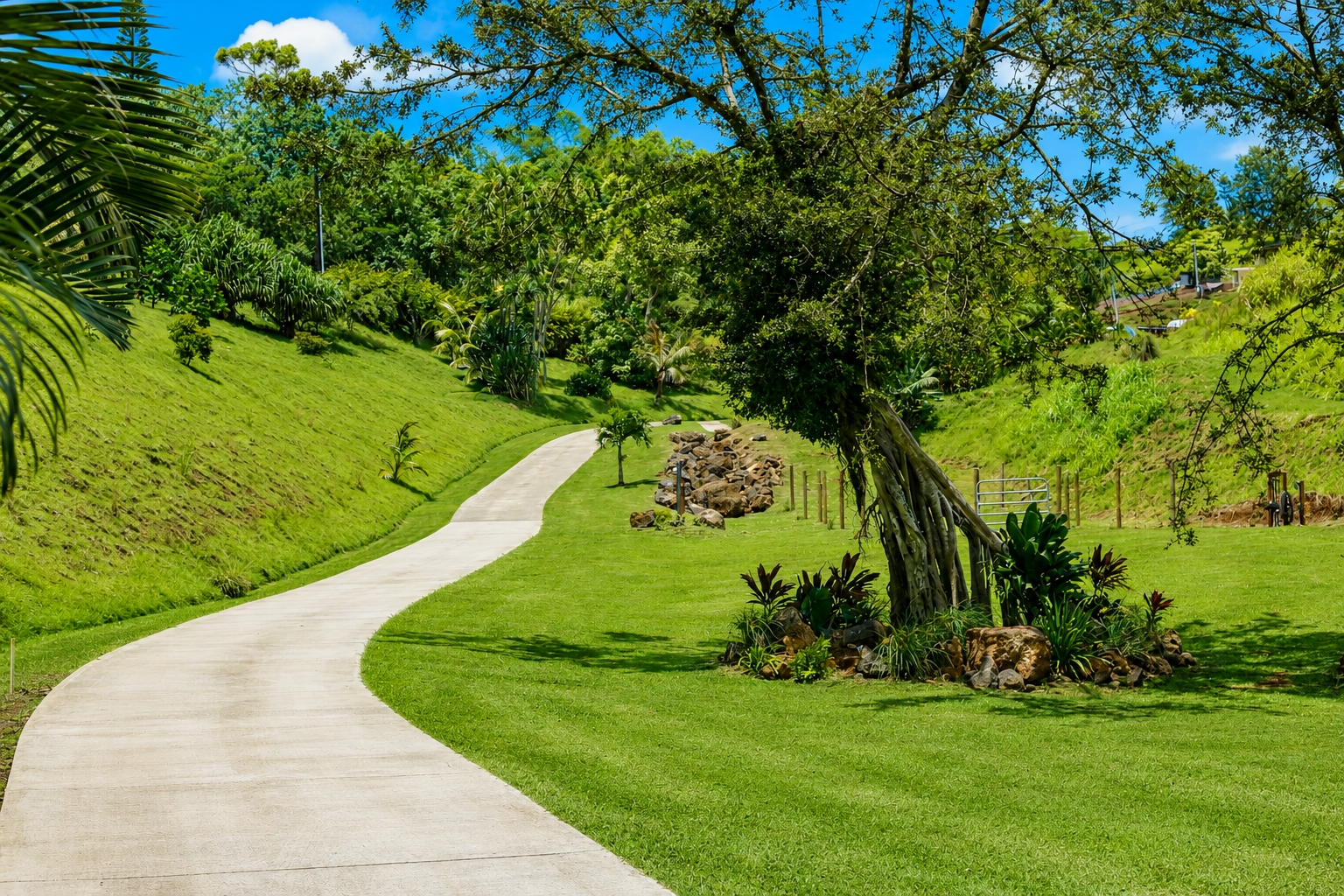 A winding concrete pathway through a lush green park with trees, grass, and decorative rocks under a blue sky.