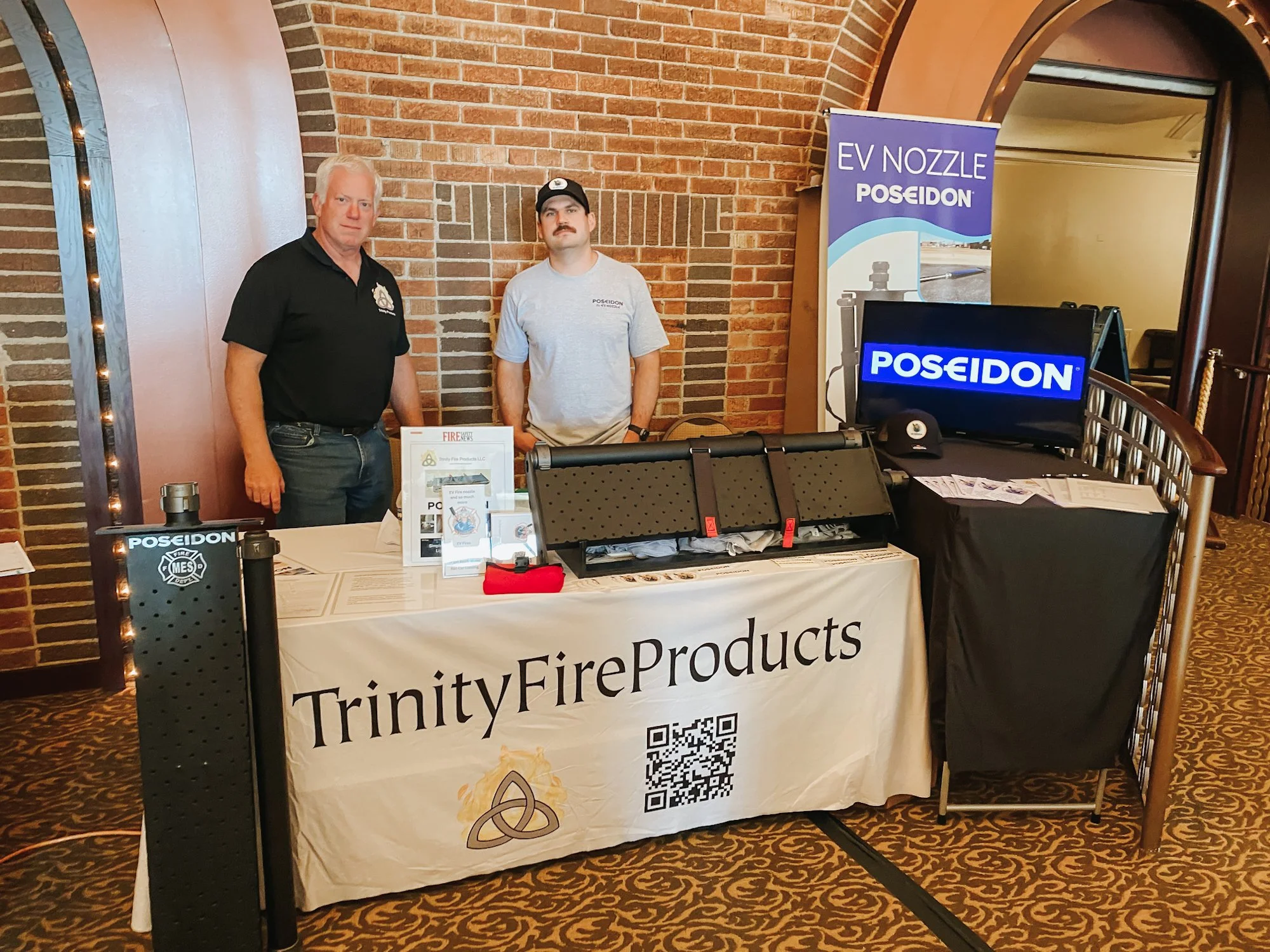 Two men standing behind a table at Trinity Fire Products booth with a large banner and digital screen. The table has informational materials, a QR code, and a small helmet. The man on the left has gray hair and is wearing a black shirt, and the man on the right has dark hair, a mustache, and is wearing a light gray shirt and cap. The background has a brick wall.