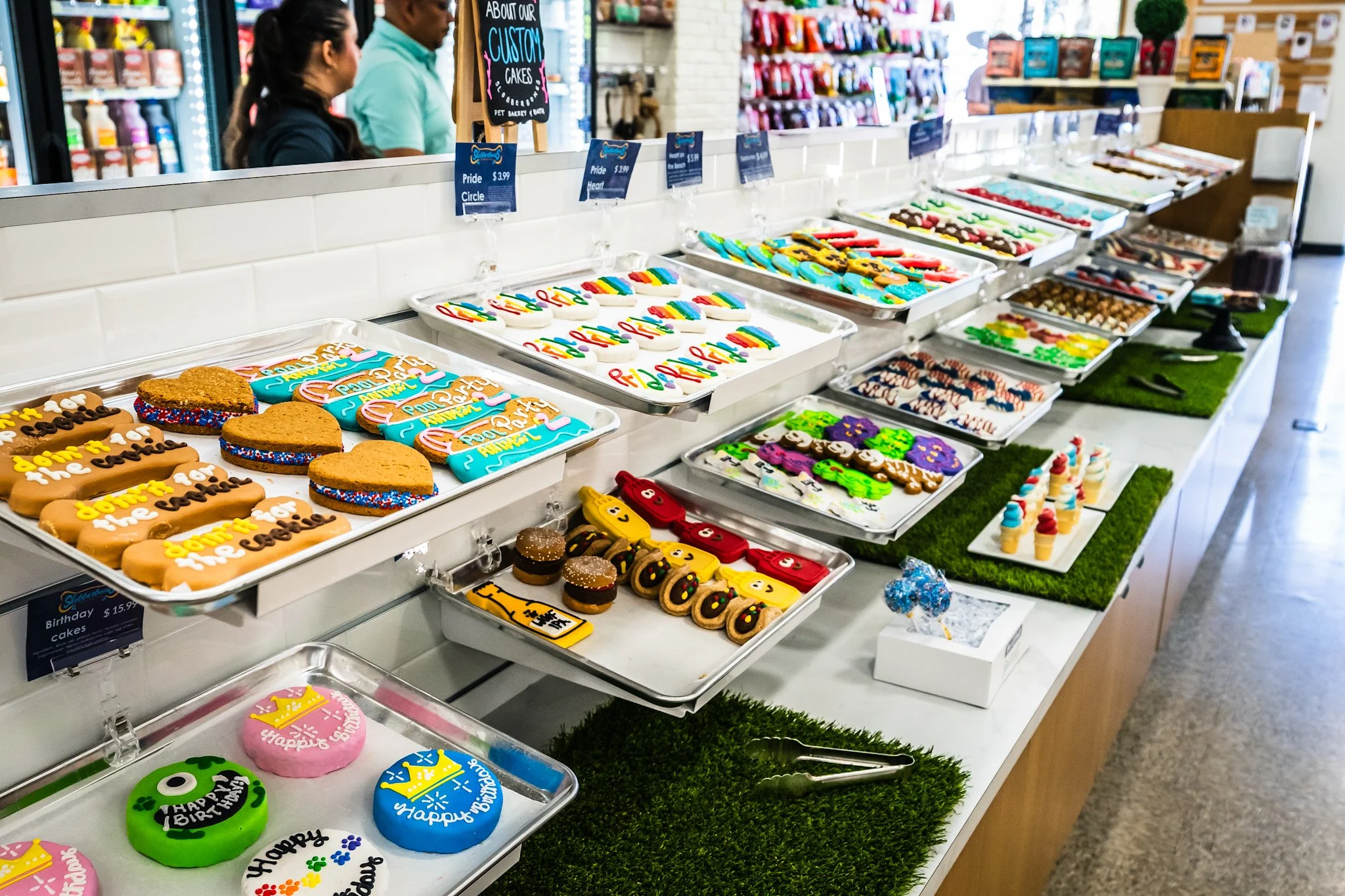 Display of decorated cookies and treats on a bakery counter inside a store, with two people shopping in the background.
