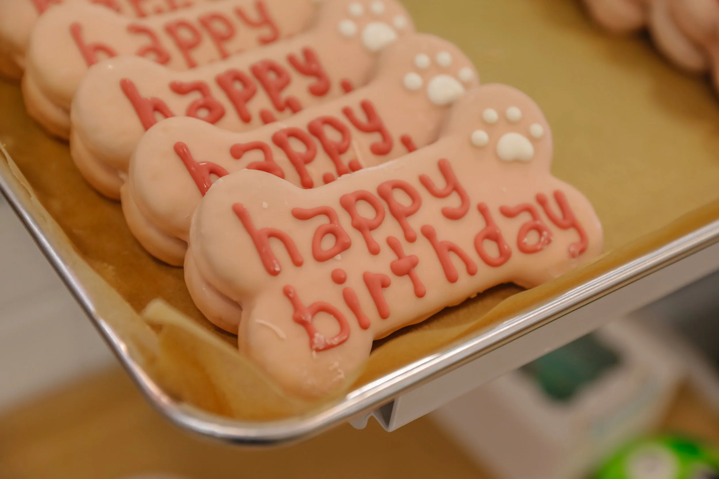 Decorative pink cookies shaped like a bone with the words 'happy birthday' written in red icing and topped with small white sugar pearls, placed on a tray.