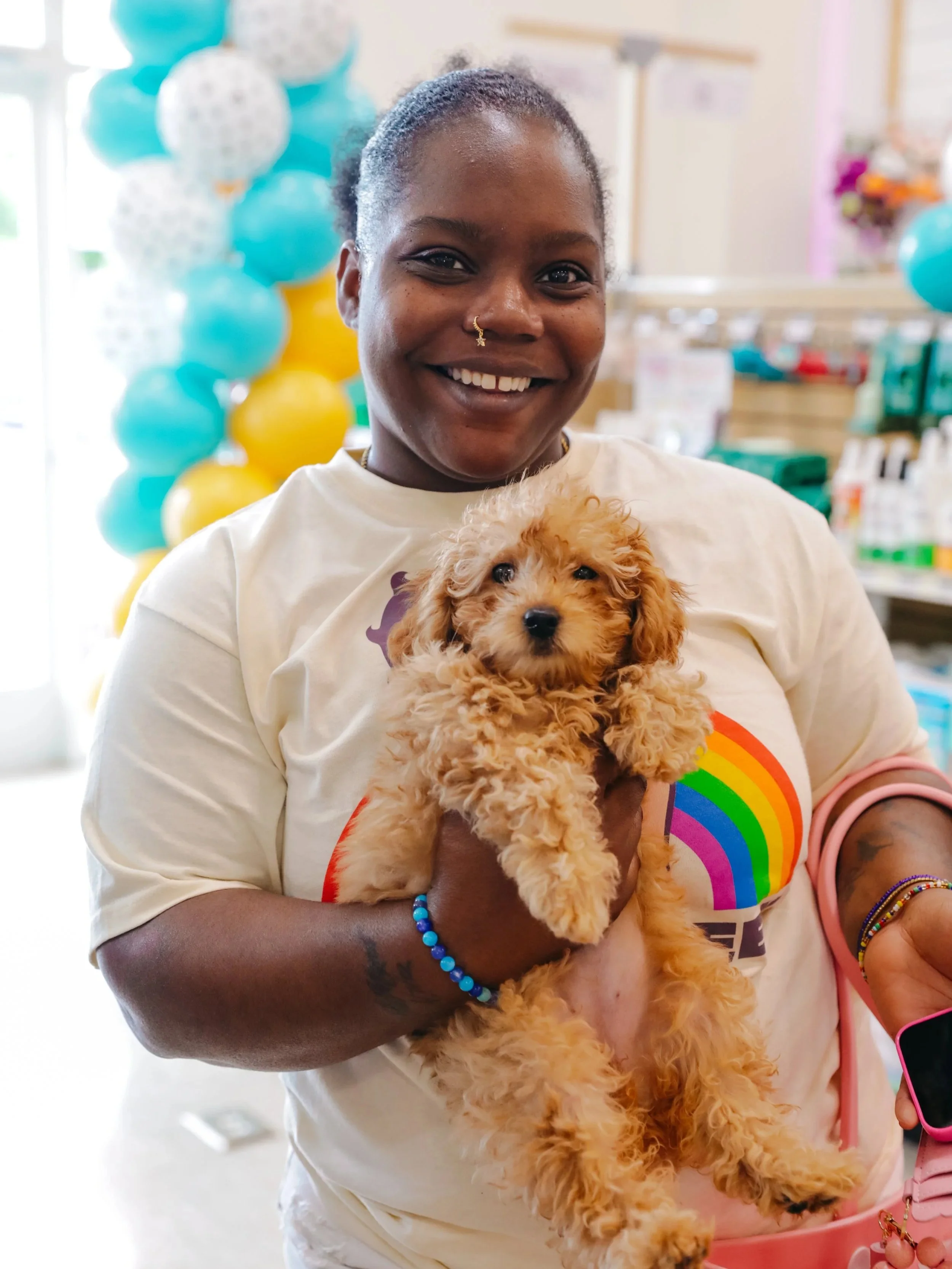A woman with dark skin and a nose piercing holding a small, fluffy, ginger-colored puppy at an indoor event with balloons in the background.