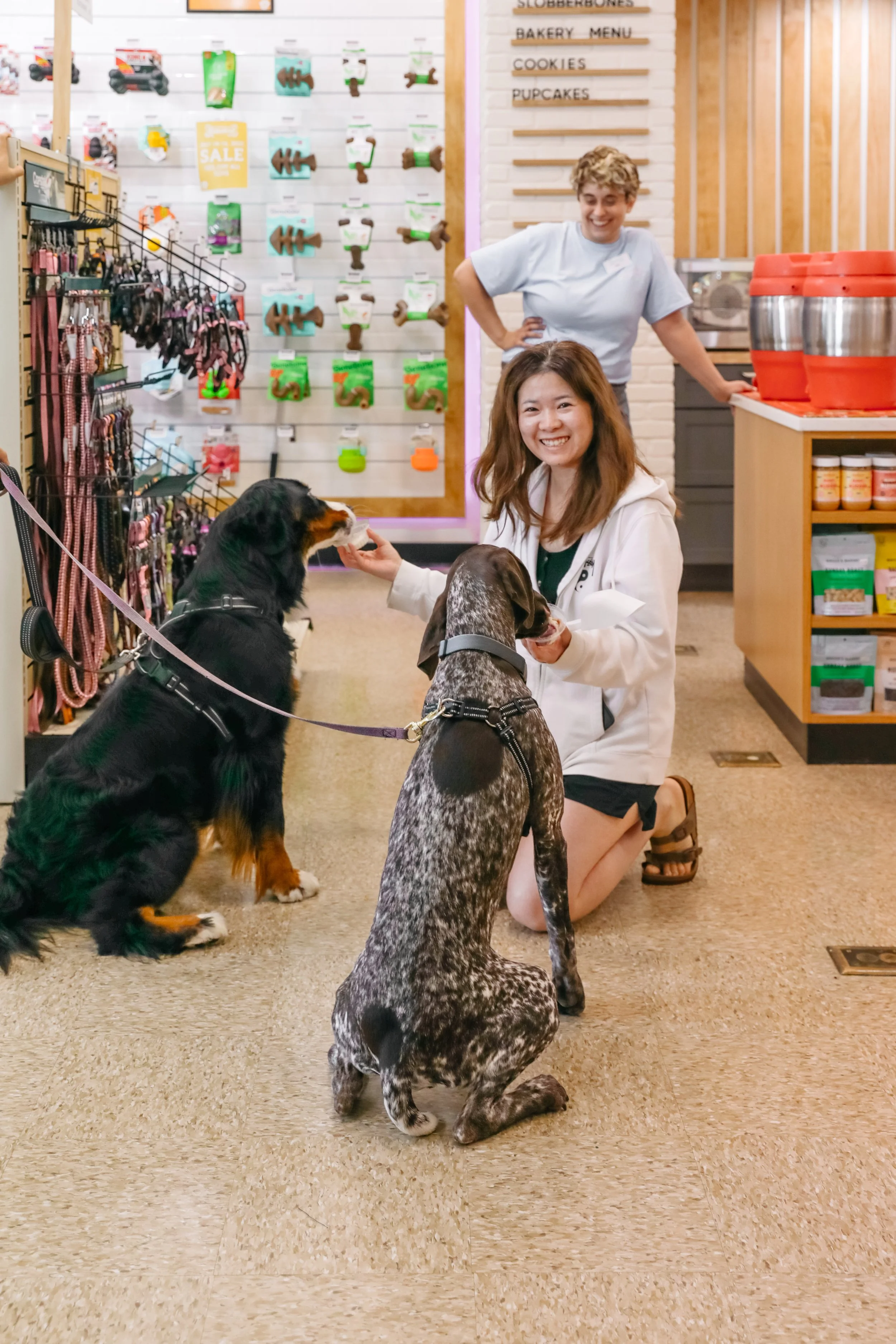 Two women and three dogs inside a pet store, with one woman kneeling and feeding a dog, and another woman standing in the background smiling.
