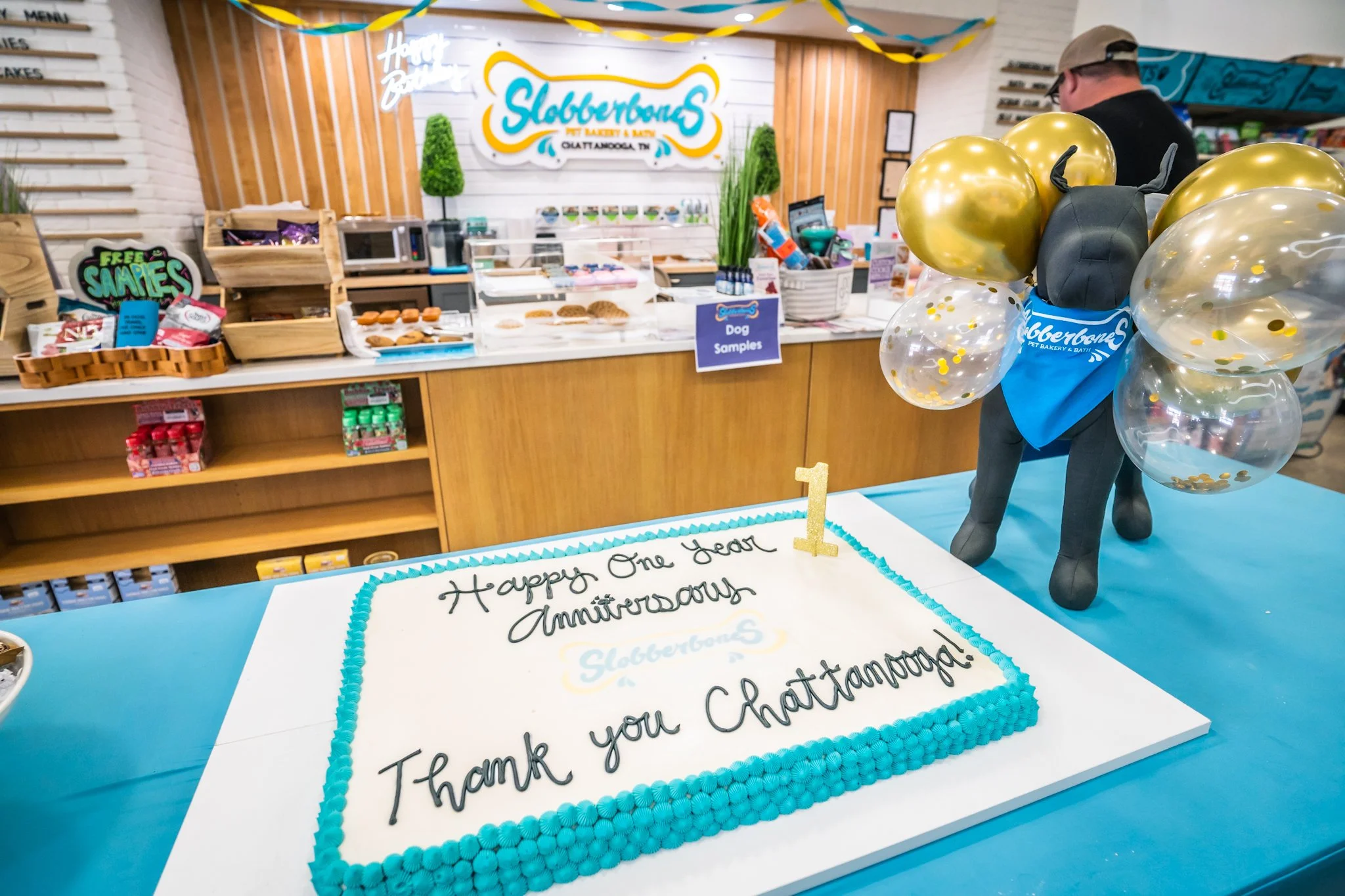A decorated bakery shop celebrating its anniversary with balloons, a cake, and a sign thanking customers. The cake has a message reading 'Happy One Year Anniversary' and is decorated with blue icing and a gold '1' topper. The bakery's sign reads 'Slu