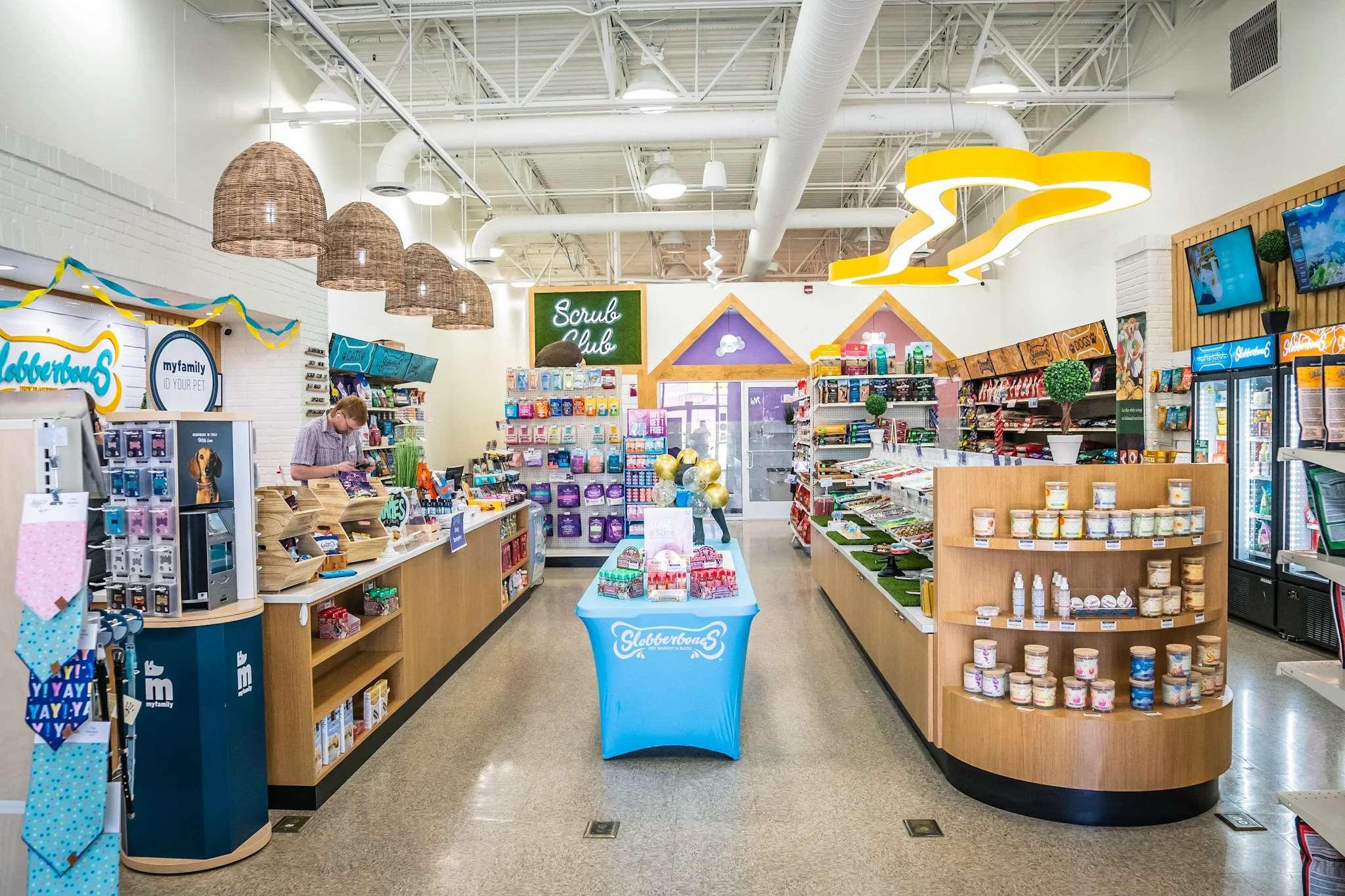 Interior of a pet store with shelves stocked with pet supplies, a checkout counter with a young man, decorative lights, and a sign for 'Scruble Club' in the background.