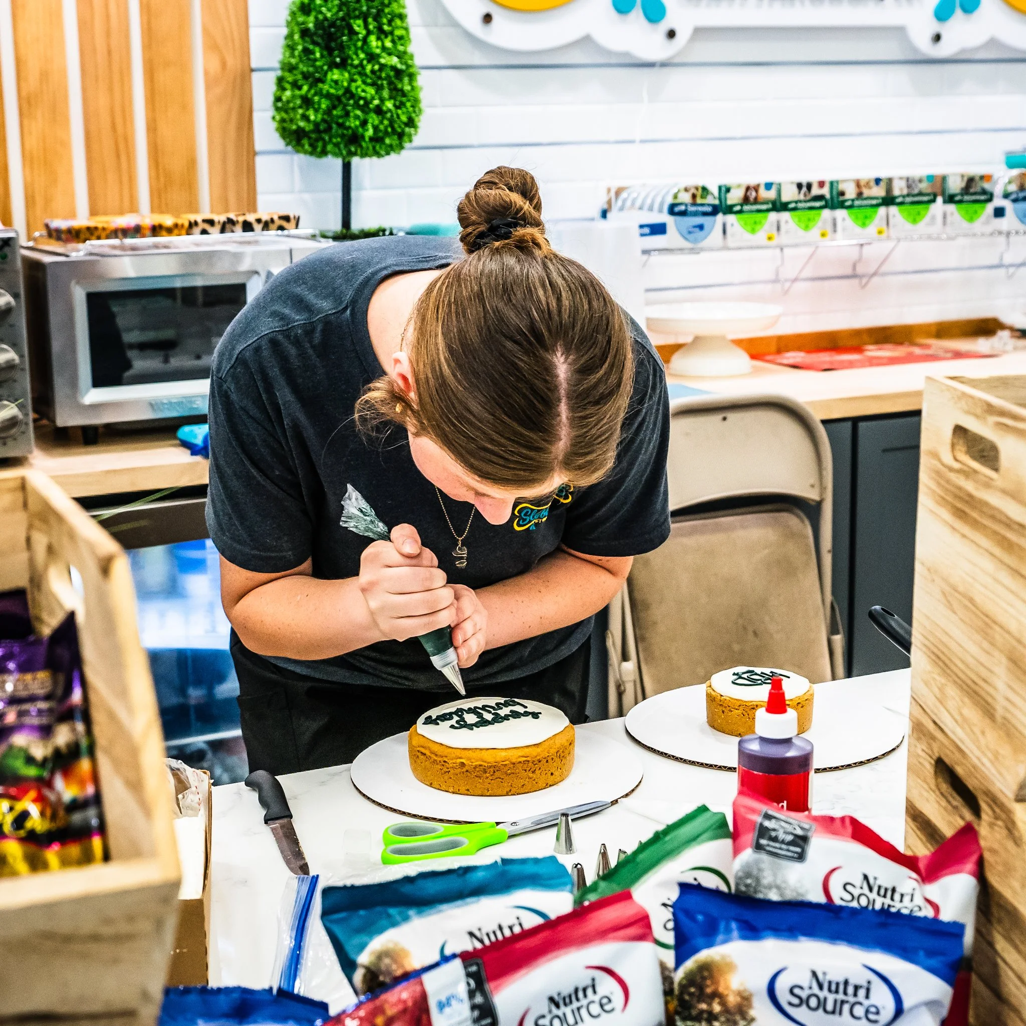 A person with brown hair tied in a bun wearing a black T-shirt is decorating a cake with black icing, possibly in a bakery or cake shop. The cake is on a table along with various tools like scissors, piping bags, and a red squeeze bottle. The backgro