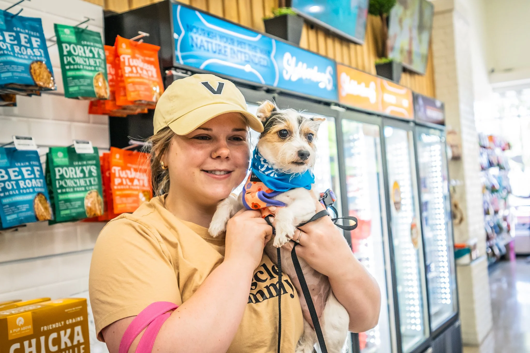 A smiling woman wearing a beige cap and tan T-shirt holding a small white and brown puppy with a blue bandana inside a pet store.