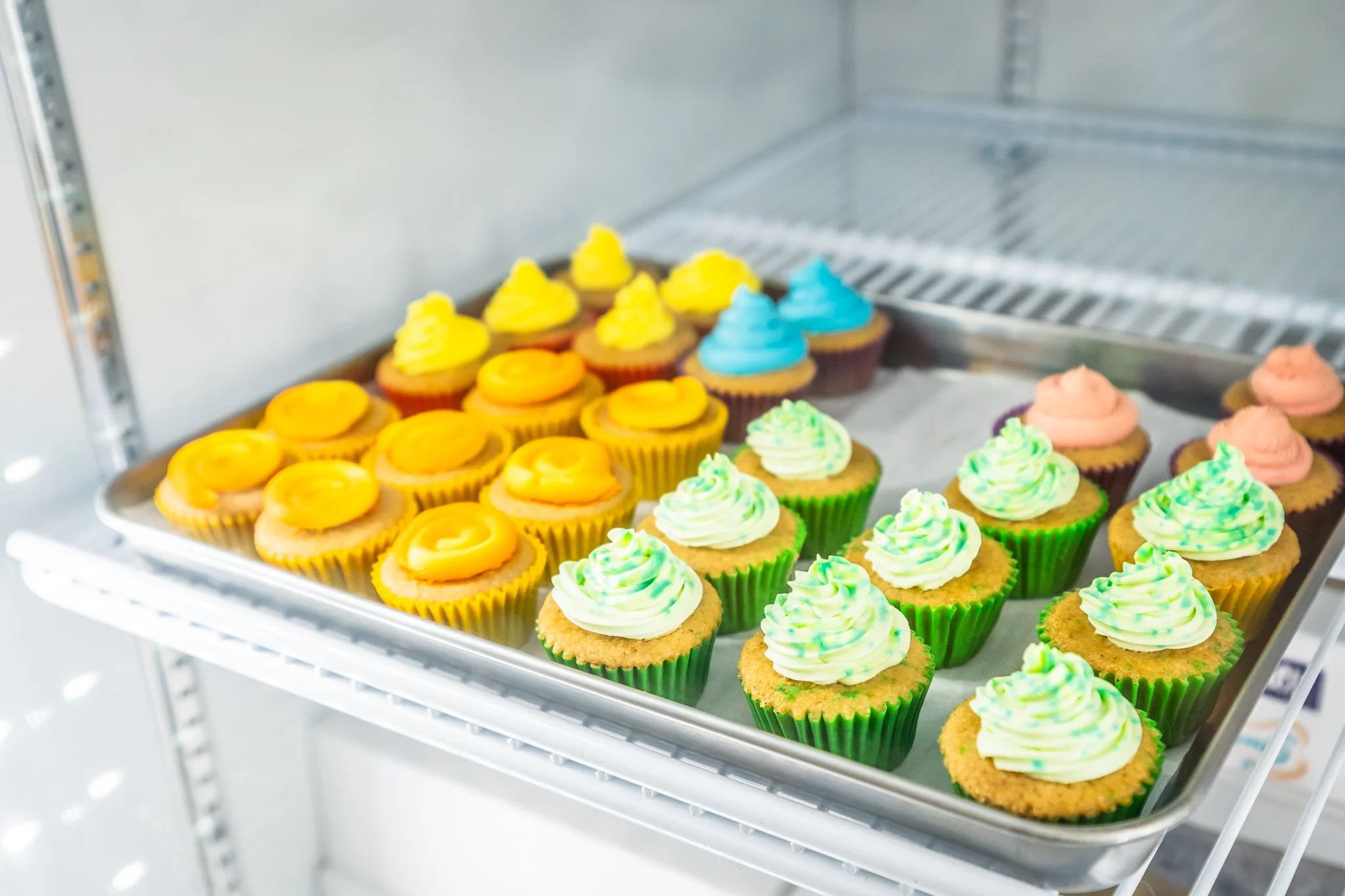 A metal baking rack with two shelves displays colorful cupcakes with various frosting designs, including yellow, blue, green, and pink, in a bright environment.