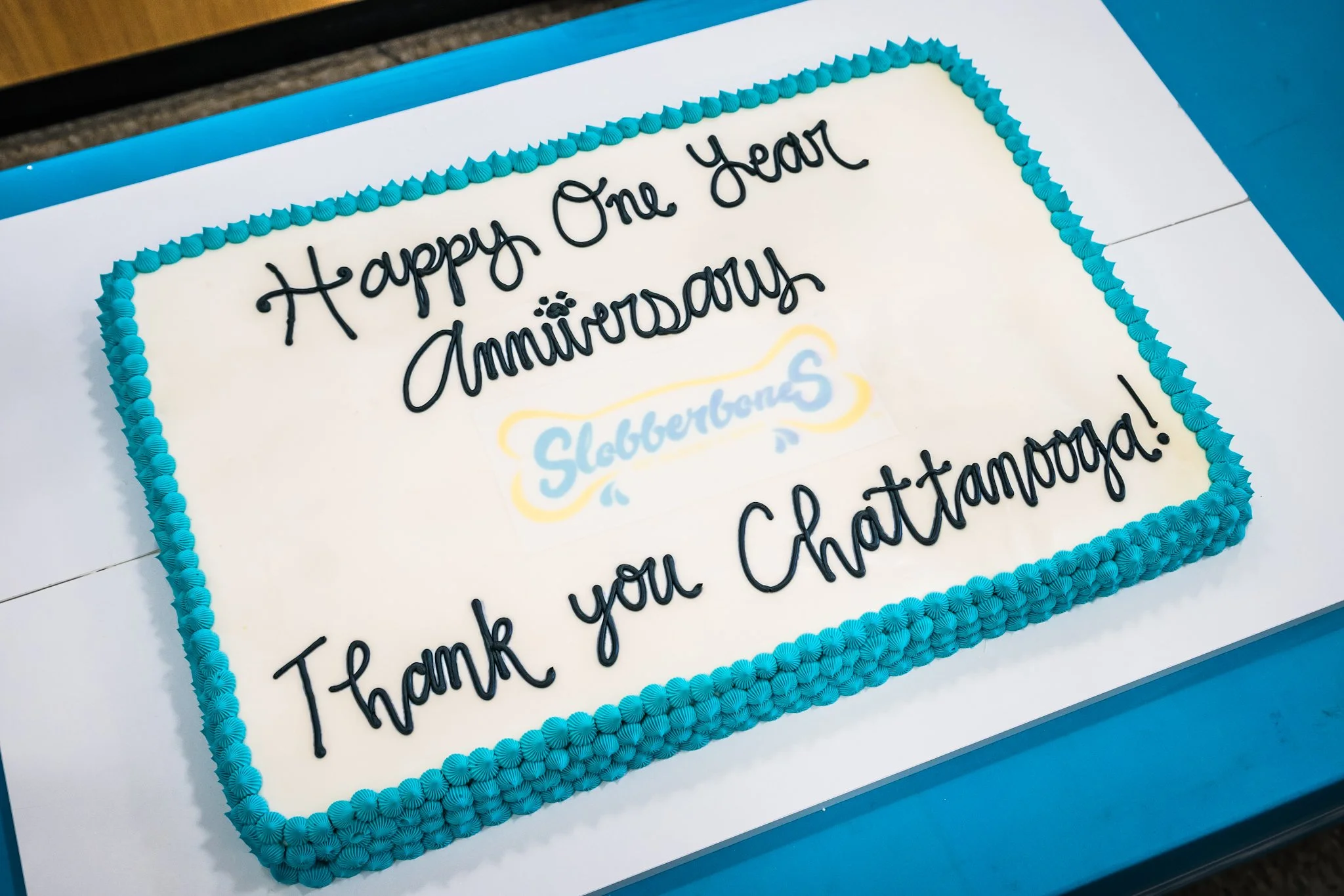 A rectangular cake with a white surface decorated with blue border piping, with the message "Happy One Year Anniversary" written in black icing and a faintly visible logo that says "Slobber Dogs."