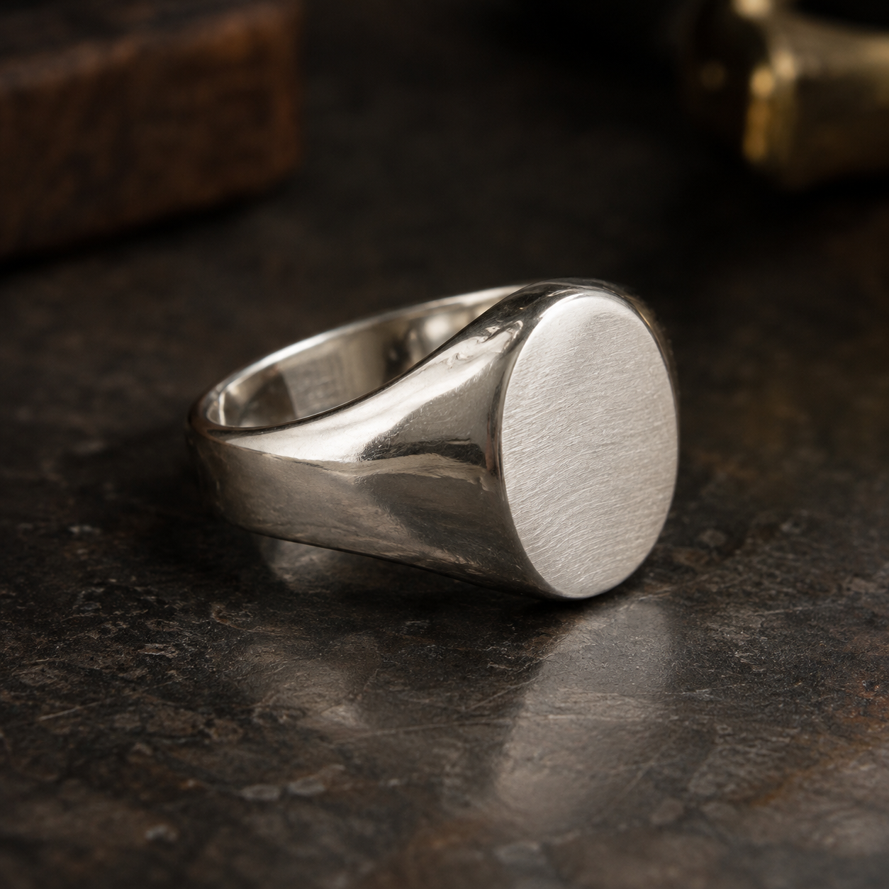 A silver signet ring with a blank, flat oval face resting on a dark, textured surface