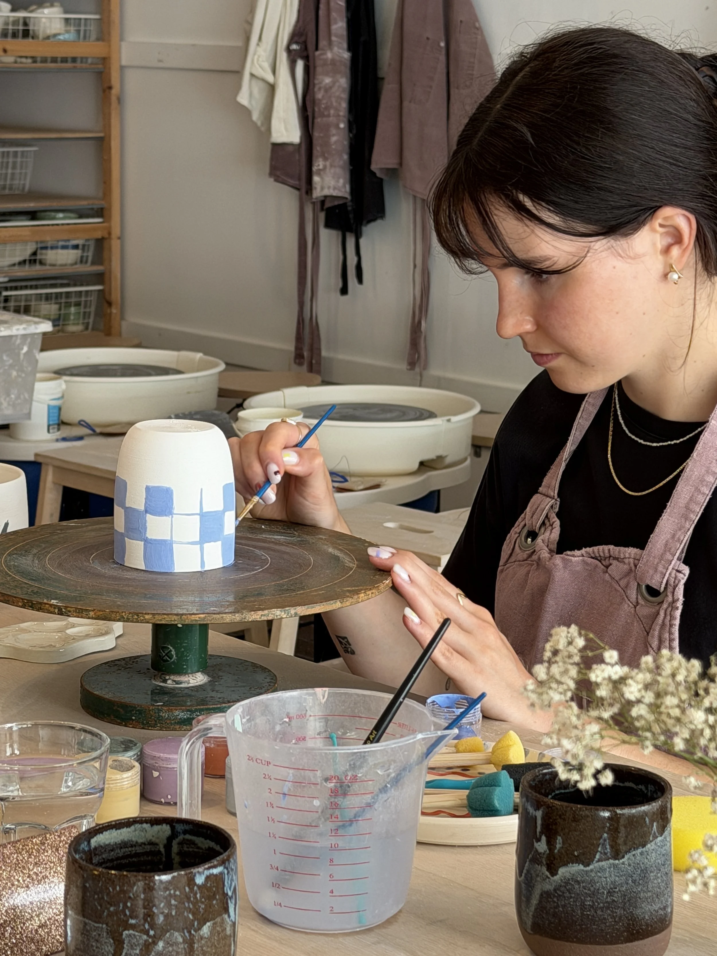 A woman painting a ceramic mug with blue and white stripes in a pottery studio. The table has various pottery supplies, including a measuring cup, painted cups, sponges, and brushes, with shelves and hanging textiles in the background.