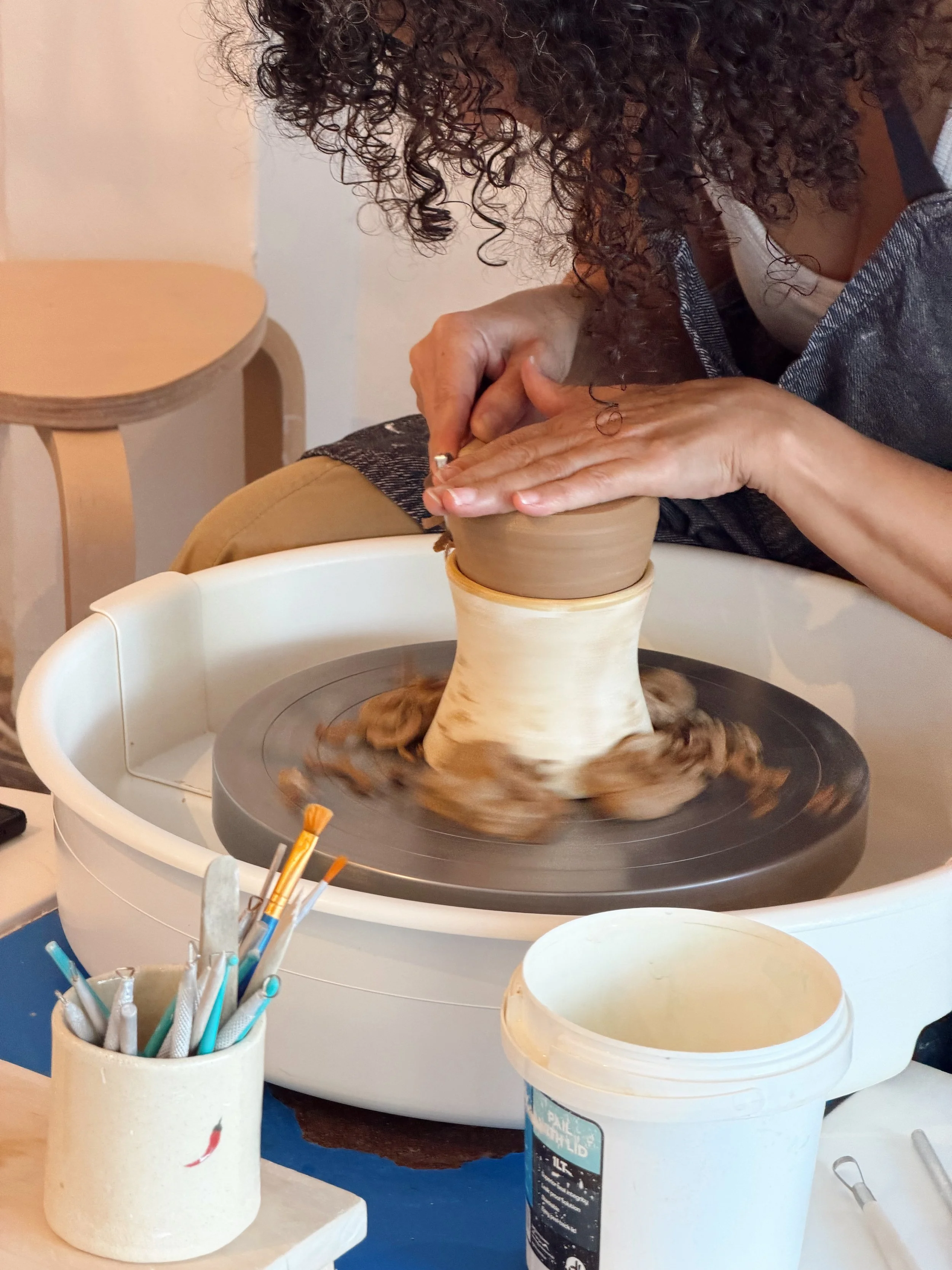 A person with curly hair making a ceramic piece on a pottery wheel, surrounded by pottery tools and supplies, in a pottery studio.