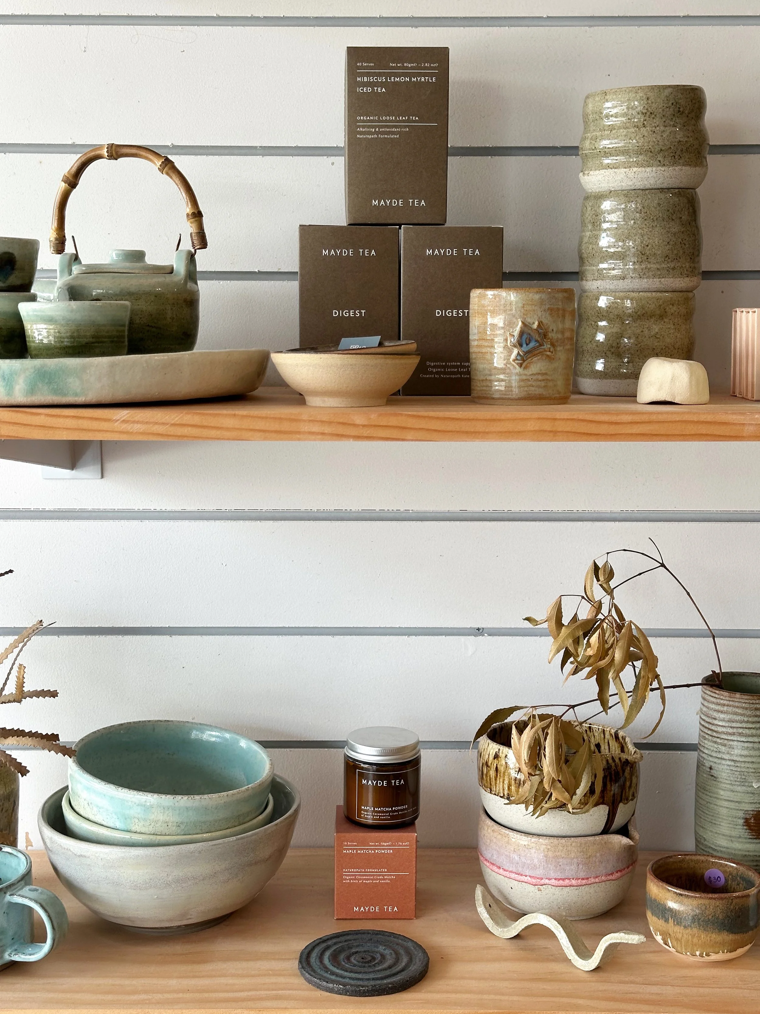 Display of ceramic and pottery items on shelves including bowls, cups, vases, and boxes, with a box of hibiscus lemon myrtle iced tea and a container of maple matcha powder in the center.
