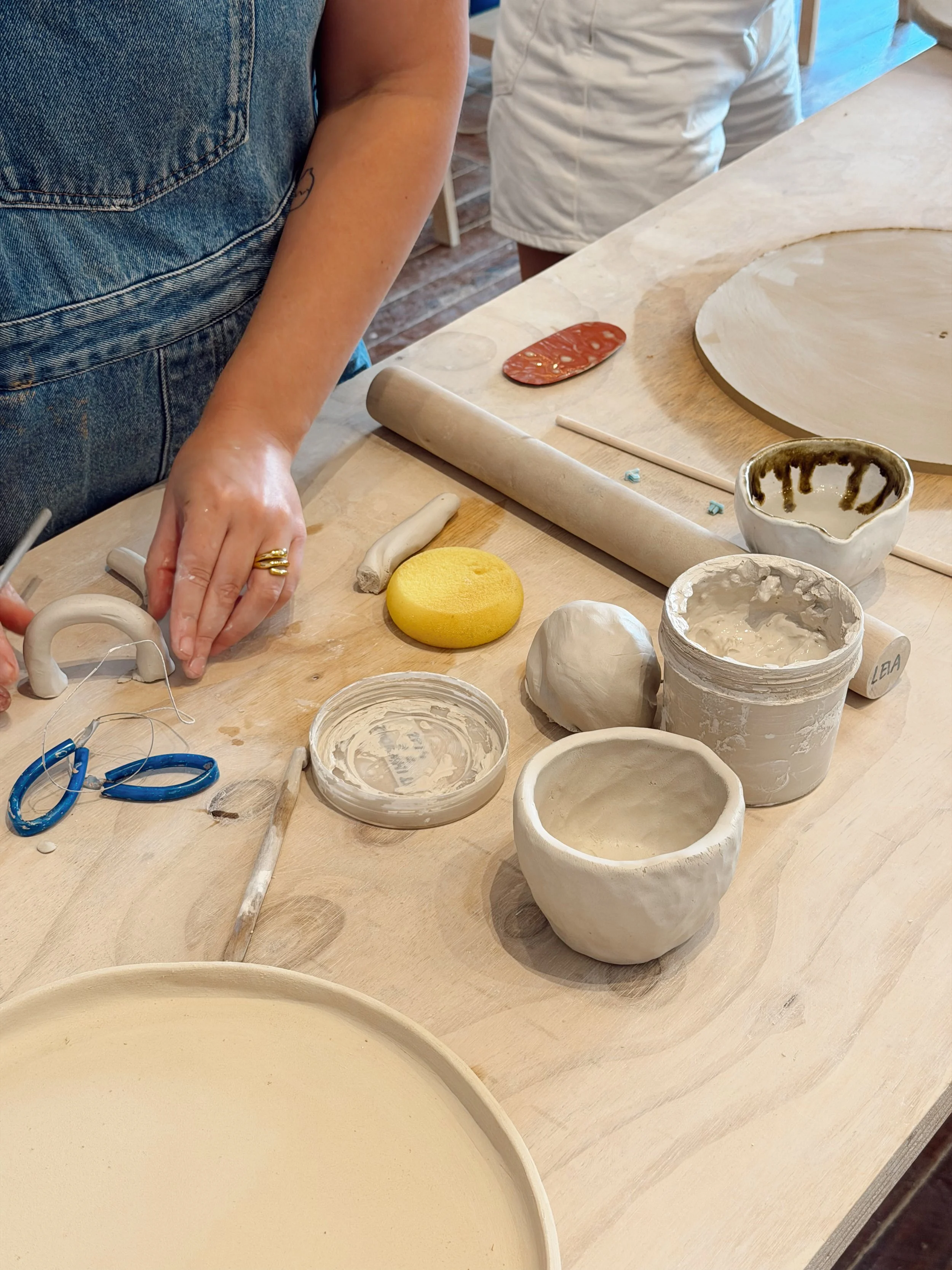 Part of a pottery or ceramics workspace showing unfinished bowls, tools, and clay pieces on a wooden table, with two people working in the background.