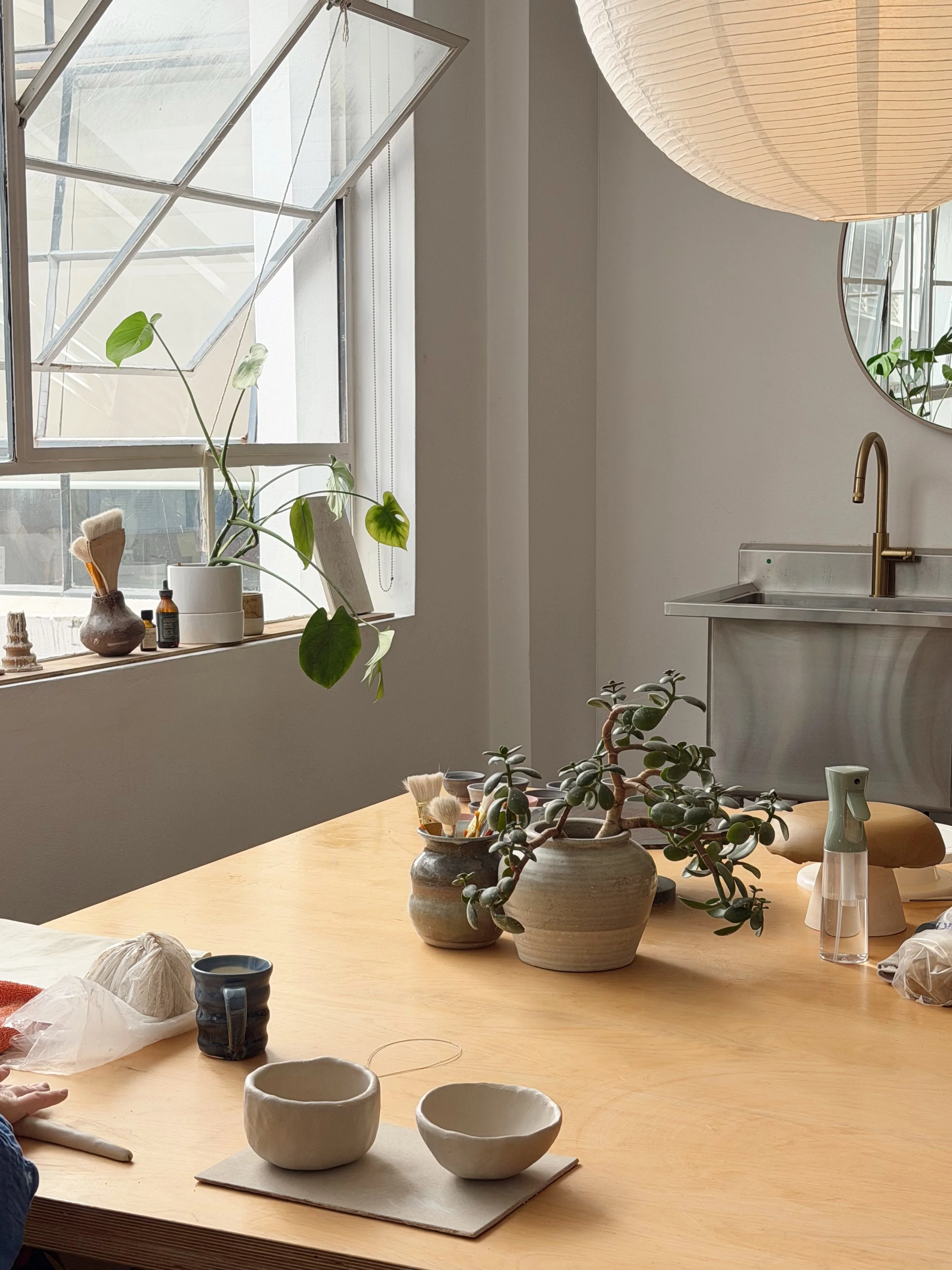 A wooden table with various unglazed ceramic bowls, a small pottery cup, and a cell phone. There are potted plants and art supplies on the table, with a window and hanging paper lantern in the background.