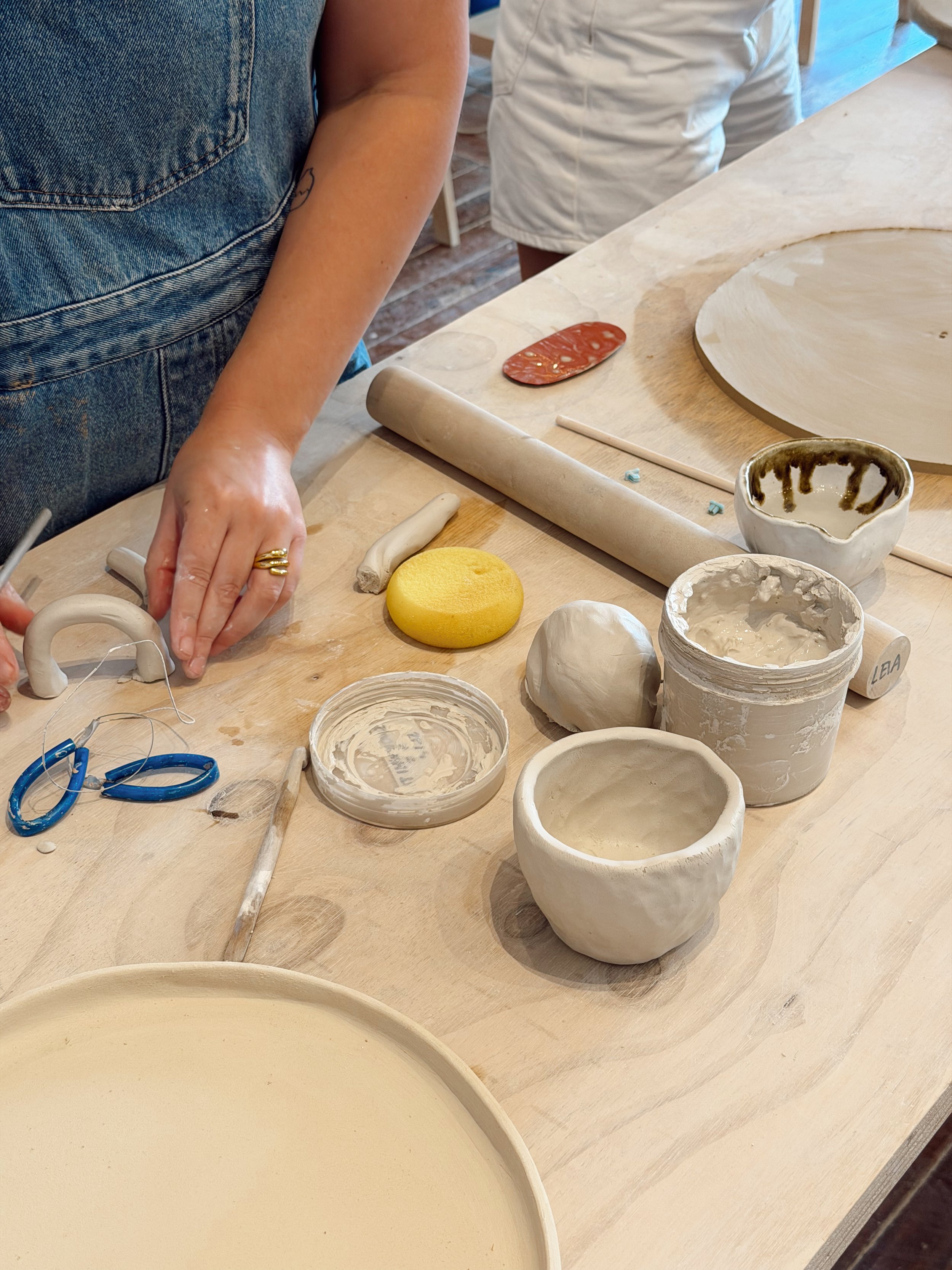People creating pottery at a workshop with various ceramic bowls, tools, and clay on the table.