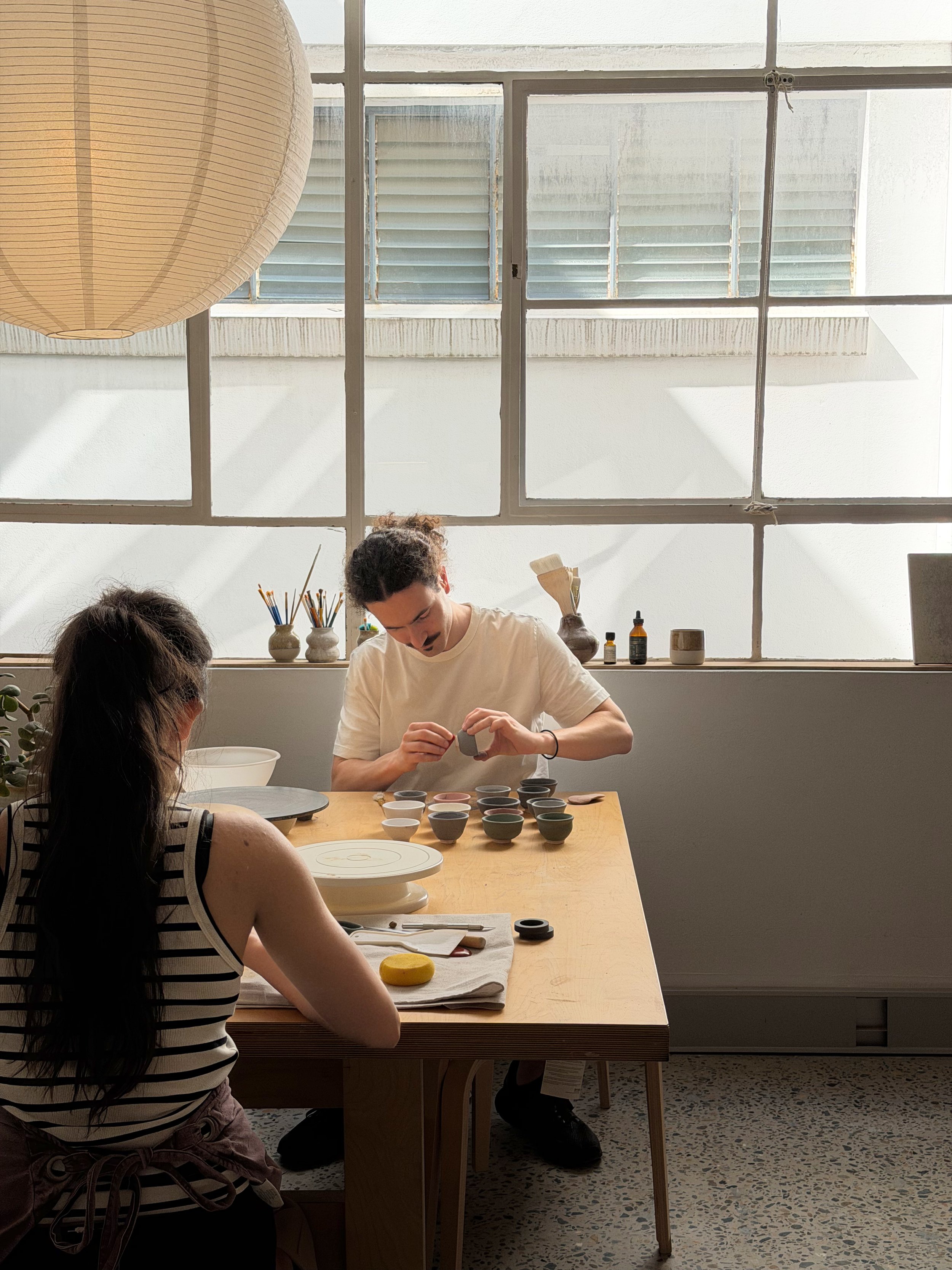 People participating in a pottery or ceramics workshop, working with small bowls and clay on a wooden table in a bright studio with large windows.