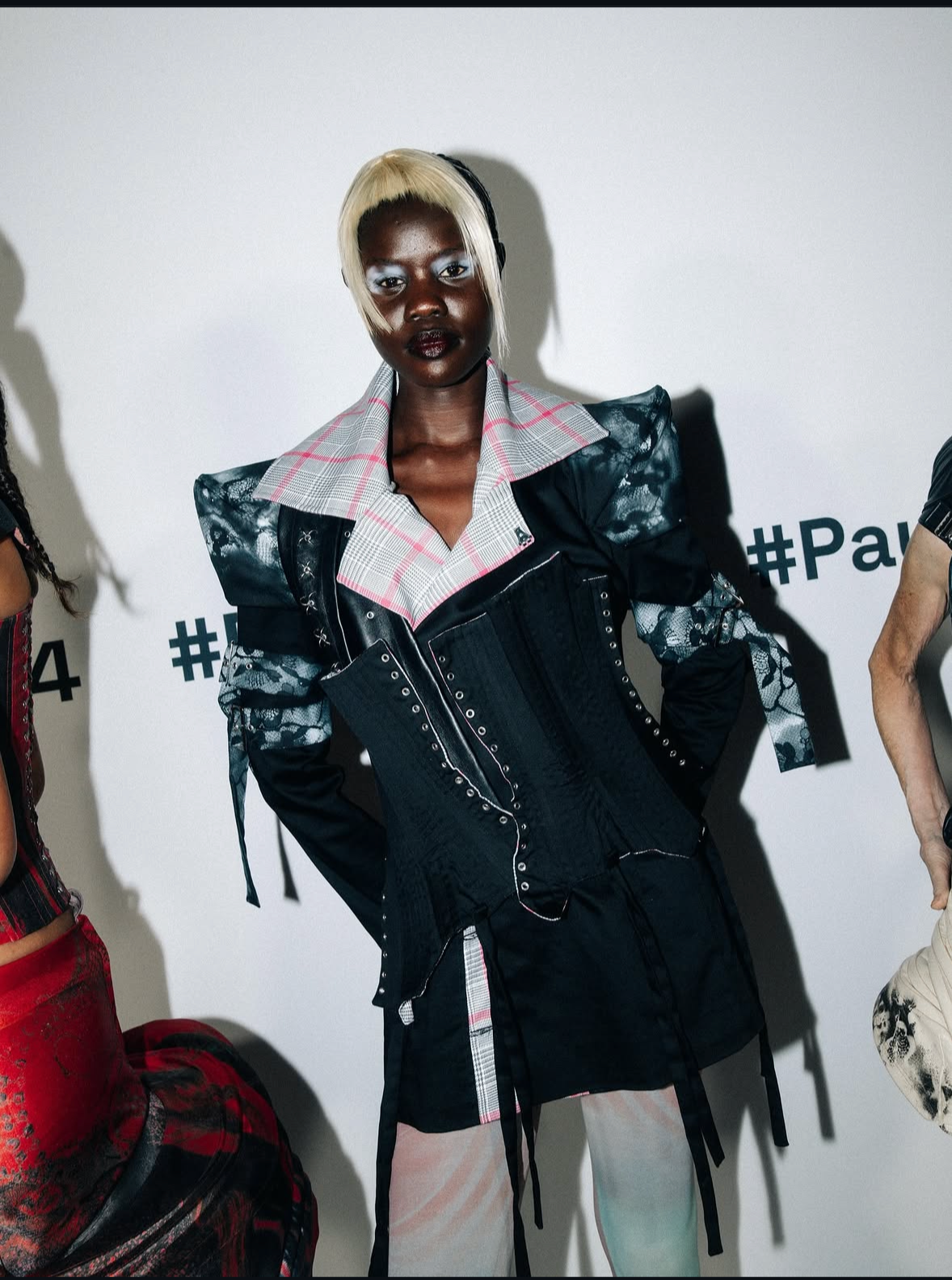 A model with dark skin and bleached blonde hair wearing a black and gray plaid jacket over a black corset and skirt, posing in front of a white wall with partially visible black text.