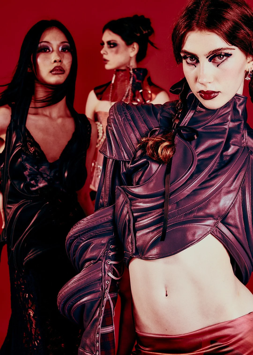 Three women with intense makeup and edgy outfits against a red background.