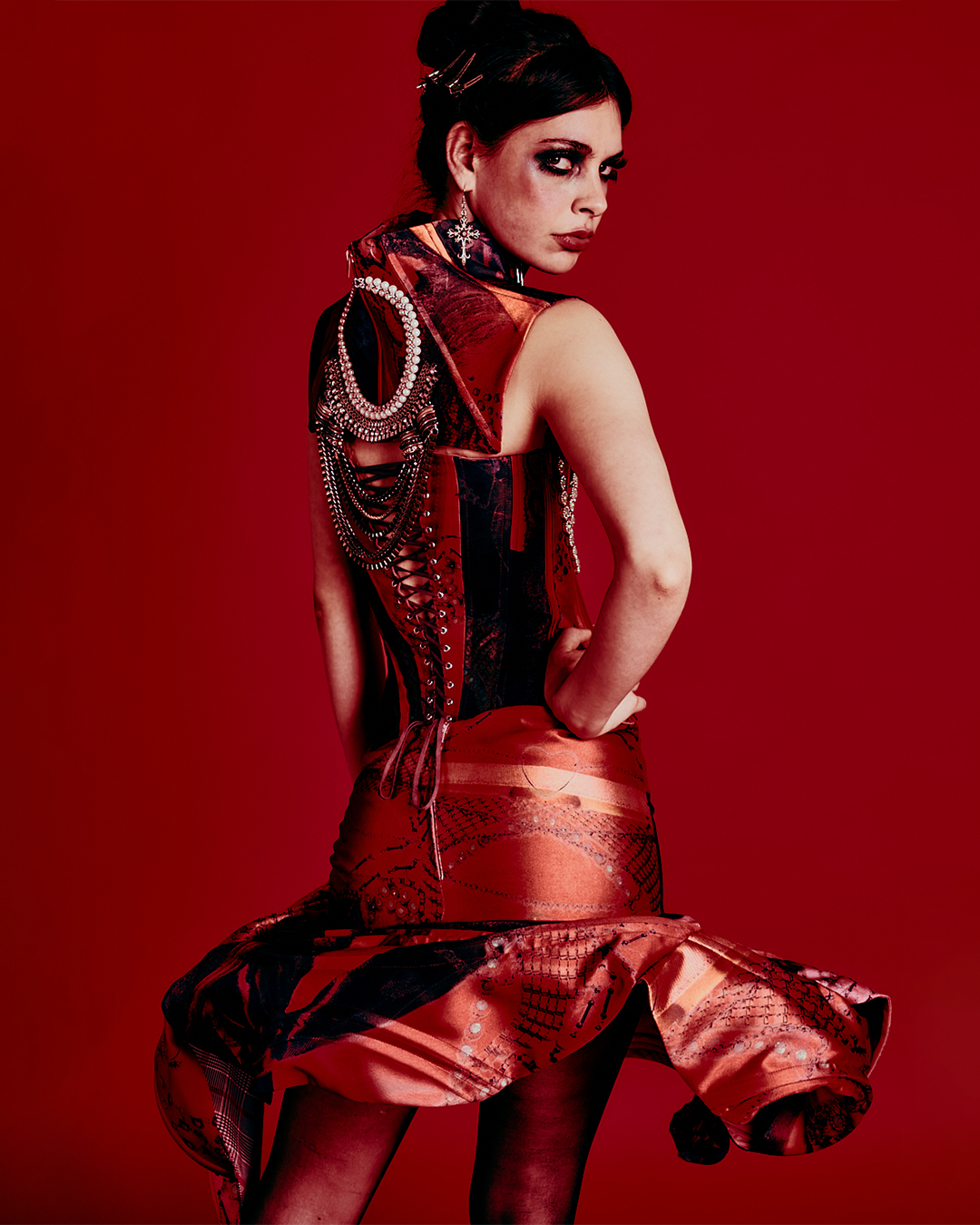 A woman with dark hair styled with clips, wearing dramatic makeup with dark eye makeup and red lipstick, poses against a solid red background. She is dressed in a red and black dress with intricate patterns and metallic accents, along with layered ch
